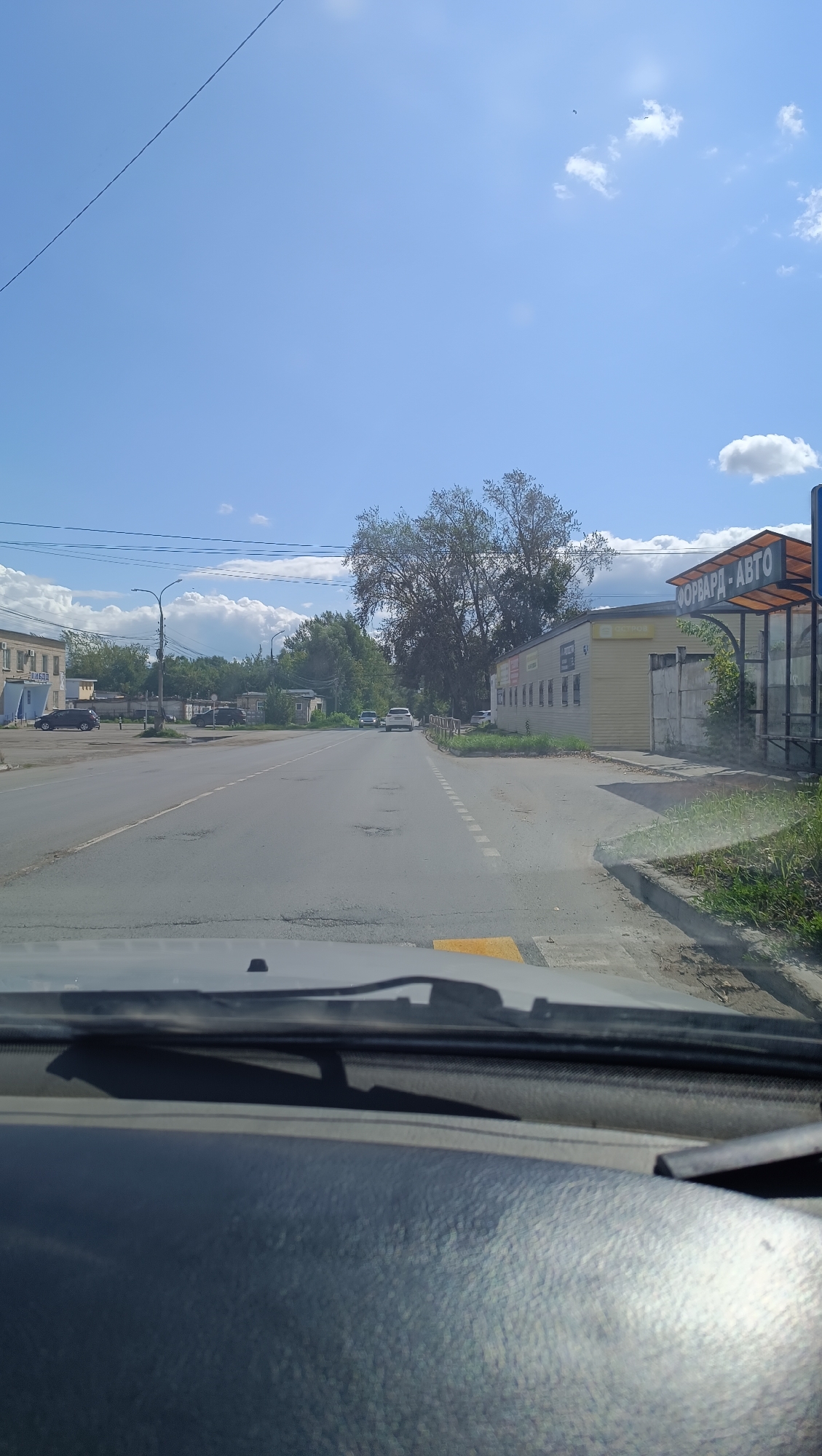 The image captures a view from inside a vehicle driving on a road in the city of Chaikovsky, Russia. The road is paved and shows signs of wear with visible potholes. On the left side of the road, there are buildings and parked cars. On the right side, a yellow building with the signage "ФОРВАРД - АВТО" (FORWARD - AUTO) is prominent, appearing to be a commercial establishment. A bus stop shelter is situated next to this building. The environment is characterized by a clear blue sky with scattered clouds and green trees lining the background. The scene is captured during daylight hours.