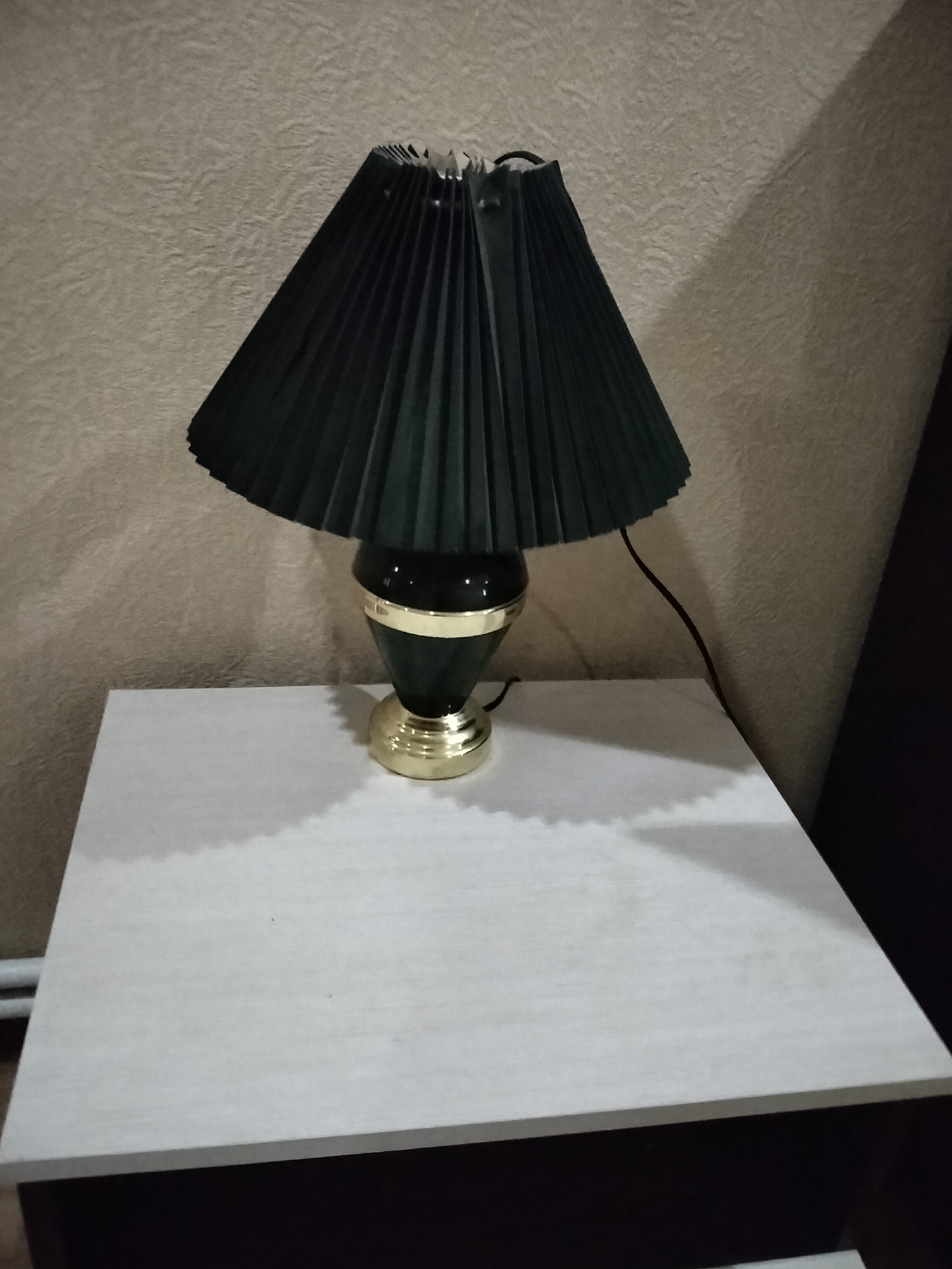A pleated, dark green lampshade rests on a gold and dark green lamp base. The lamp sits on a light-colored, textured surface. Behind the lamp, a textured beige wall is visible, with a shadow cast upon it from the lamp. A black power cord snakes away from the lamp base. The scene is indoors, likely in a residential setting, though no specific location in Nizhnedonskoy, Russia, can be identified from the image alone.