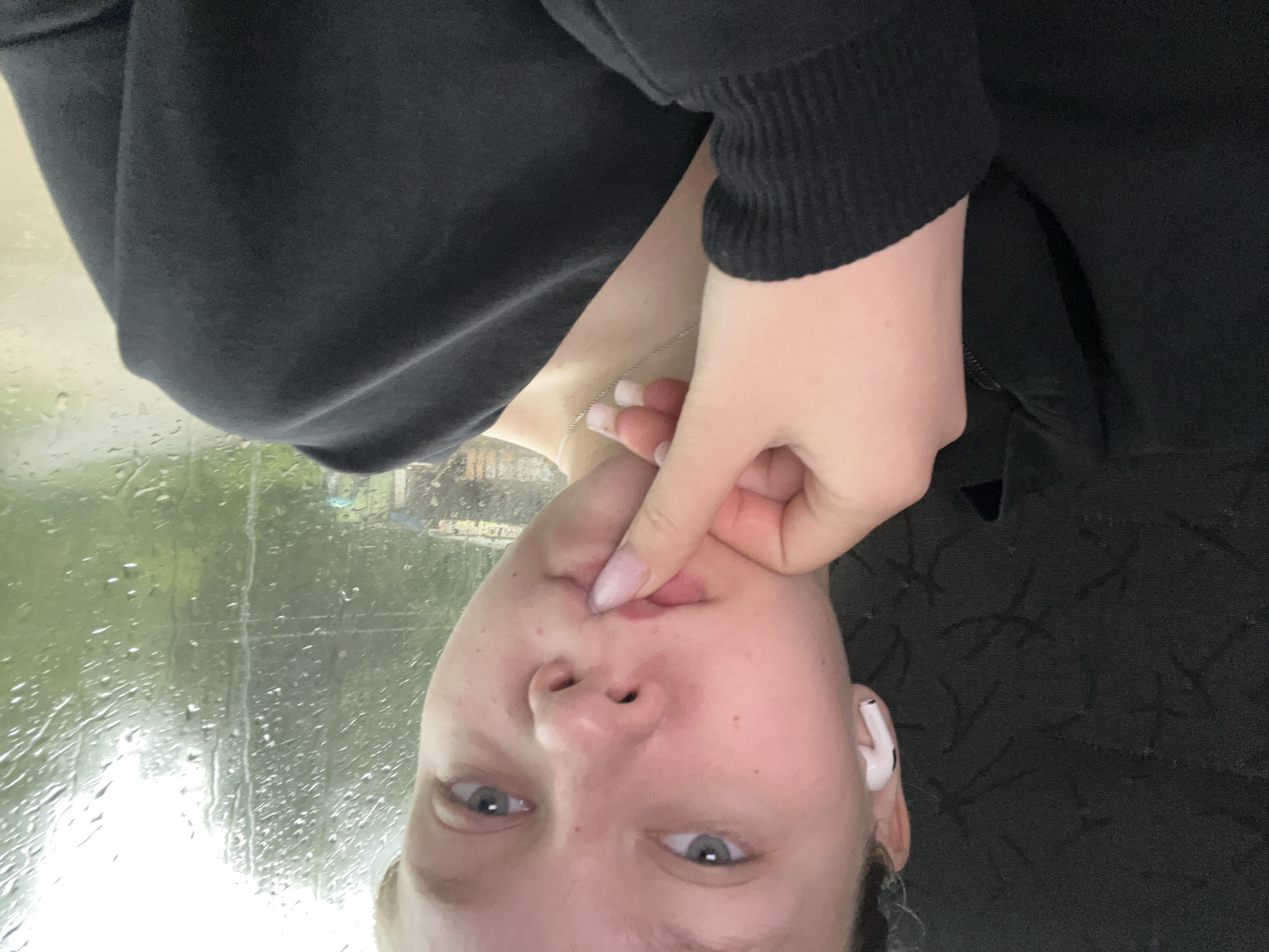 A young woman with light skin and light blue eyes is pictured in a close-up, waist-up shot. She is wearing a black sweatshirt and white earbuds in her left ear. Her right hand is raised, with her index finger touching her lips. Her nails are light pink. In the background, water droplets streak down a window, obscuring a blurry view of trees and a small building, suggesting a rainy day. The scene appears to be indoors, possibly in a vehicle, given the reflection on the window. There is no discernible information in the image to place the location in Krasnokamsk, Russia.