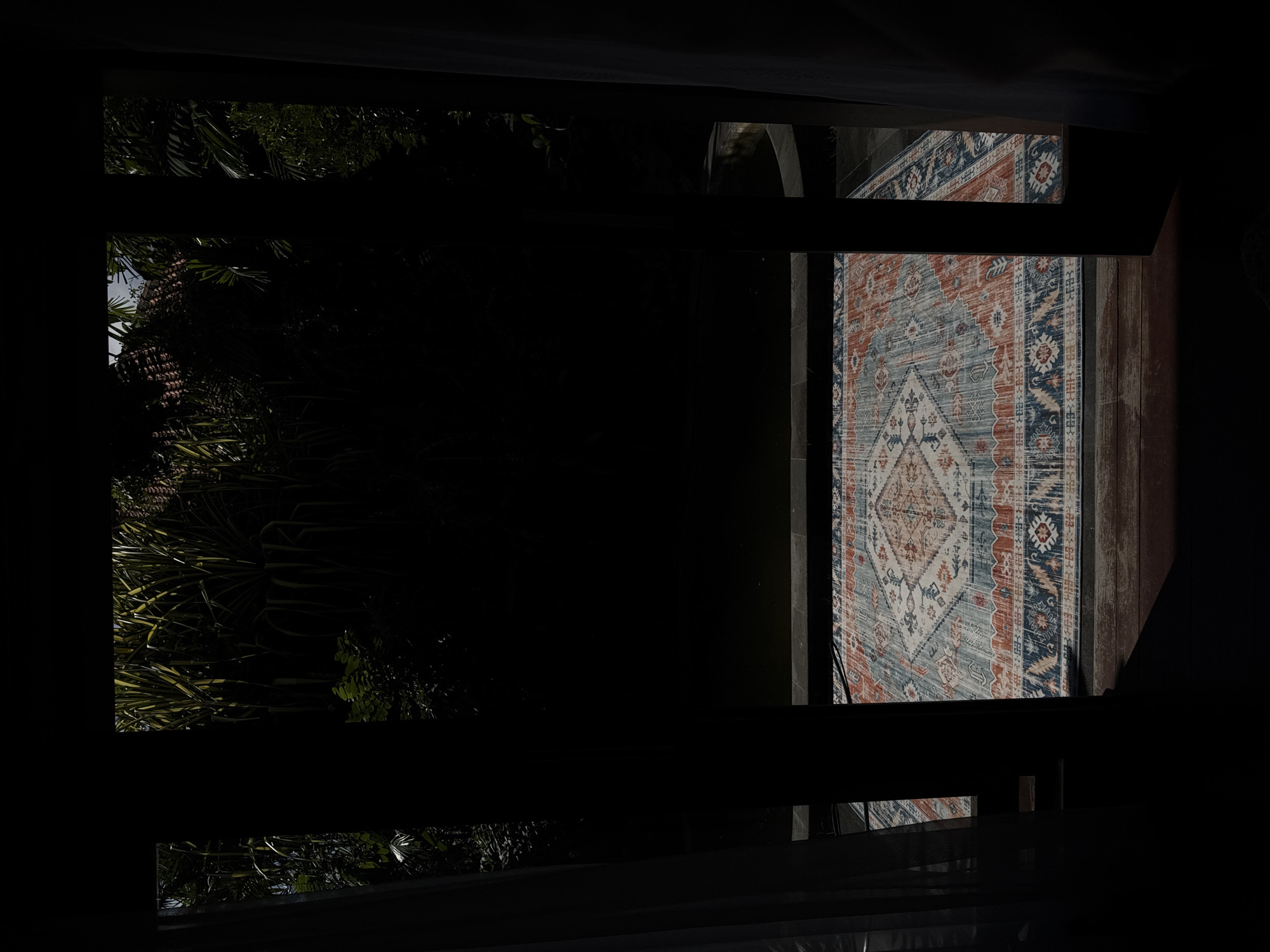 The image depicts a view through an open doorway, looking out onto an outdoor area with a patterned rug laid on a wooden deck. The rug features intricate geometric designs in shades of blue, orange, and cream. Beyond the rug, there appears to be a dark body of water, possibly a pool or pond, and lush green foliage from tropical plants. The scene is dimly lit, with strong shadows cast by the doorway frame and curtains. The setting is likely a resort or private residence in Kuta Selatan, Indonesia, given the tropical vegetation and architectural style. There are no people visible in the image.