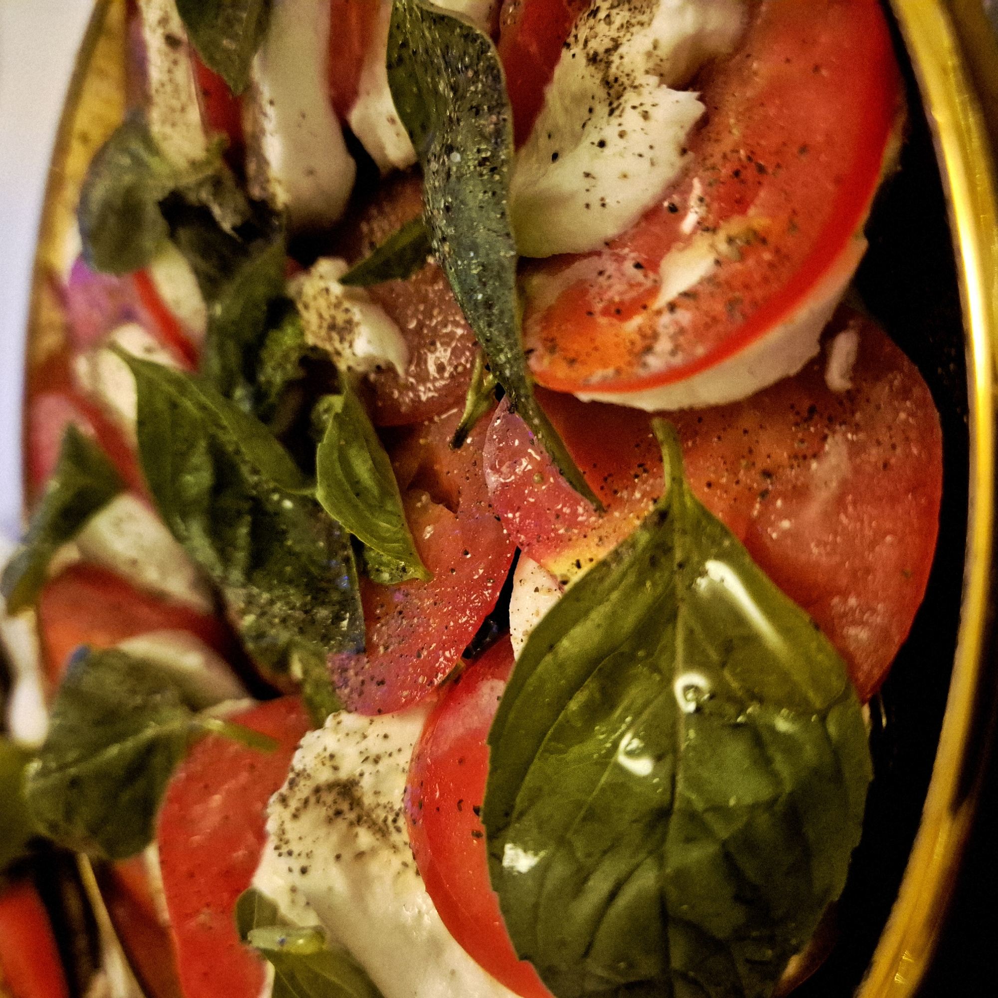 The image shows a close-up of a Caprese salad. Slices of red tomatoes and white mozzarella cheese are layered with green basil leaves. The salad is seasoned with black pepper. The salad is served in a golden bowl. The setting is indoors.