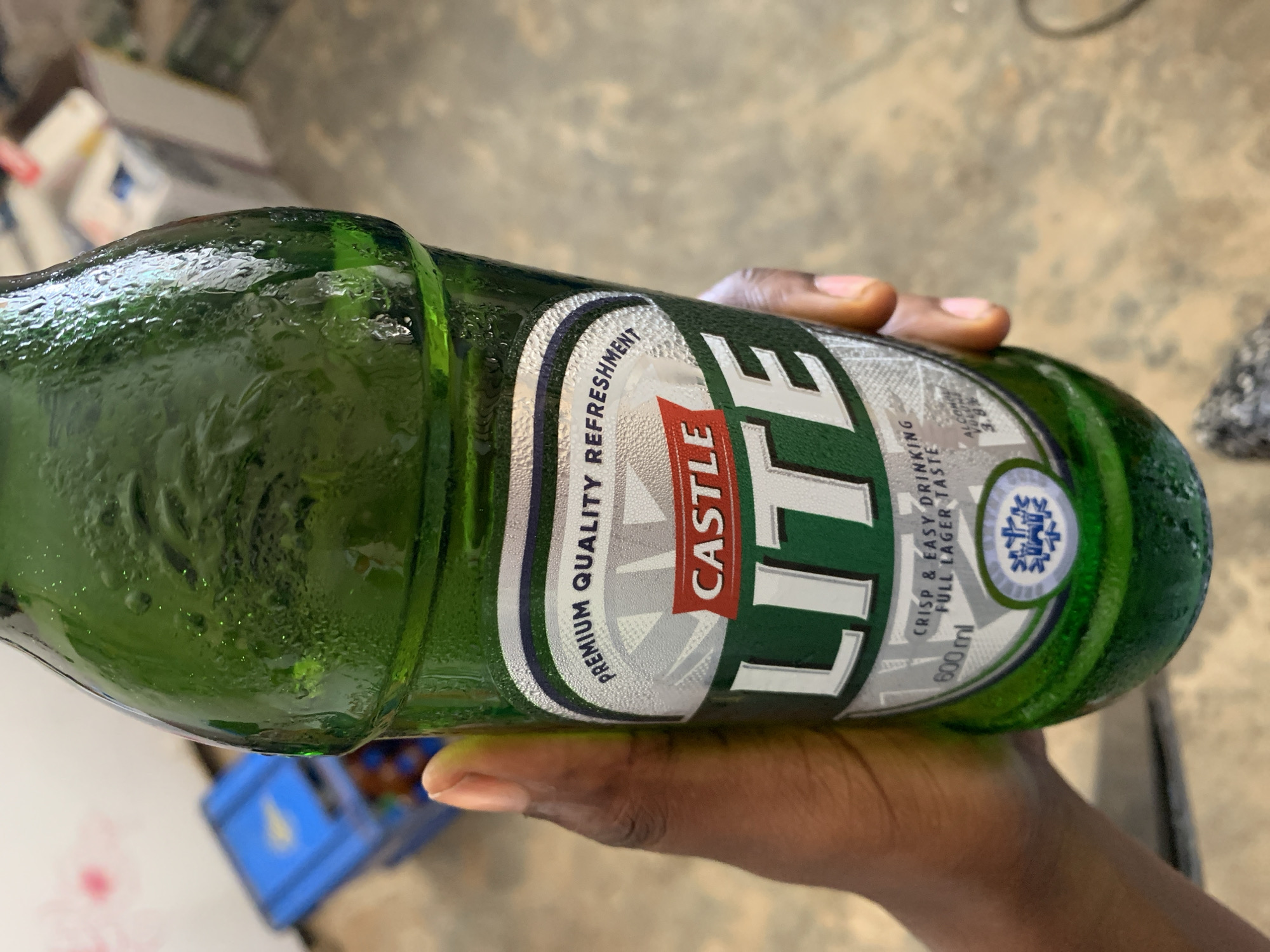 An close-up, eye-level shot captures a person's hand holding a cold green glass bottle of Castle Lite beer. The bottle is angled diagonally, with its base to the lower right and neck to the upper left. Abundant condensation and water droplets cover the entire surface of the bottle, indicating it is chilled.
The label, prominently displayed in the center, is white and green with red accents. Large white text reads "CASTLE LITE." Above "CASTLE," smaller text says "PREMIUM QUALITY REFRESHMENT." To the right of "LITE," in a vertical orientation, the label states "CRISP & EASY DRINKING" and "FULL LAGER TASTE." Below this, "600ml" is clearly visible, along with "ALC/VOL 5.0%" below that. A circular logo featuring a snowflake-like design is also present on the label. The hand holding the bottle has dark skin.
The background is out of focus, showing blurry shapes of a concrete floor and some indistinct objects, including a blue plastic crate in the lower left, hinting at an indoor or semi-enclosed setting. The scene depicts a casual moment of holding a refreshing beverage in New Stadium, Nigeria.