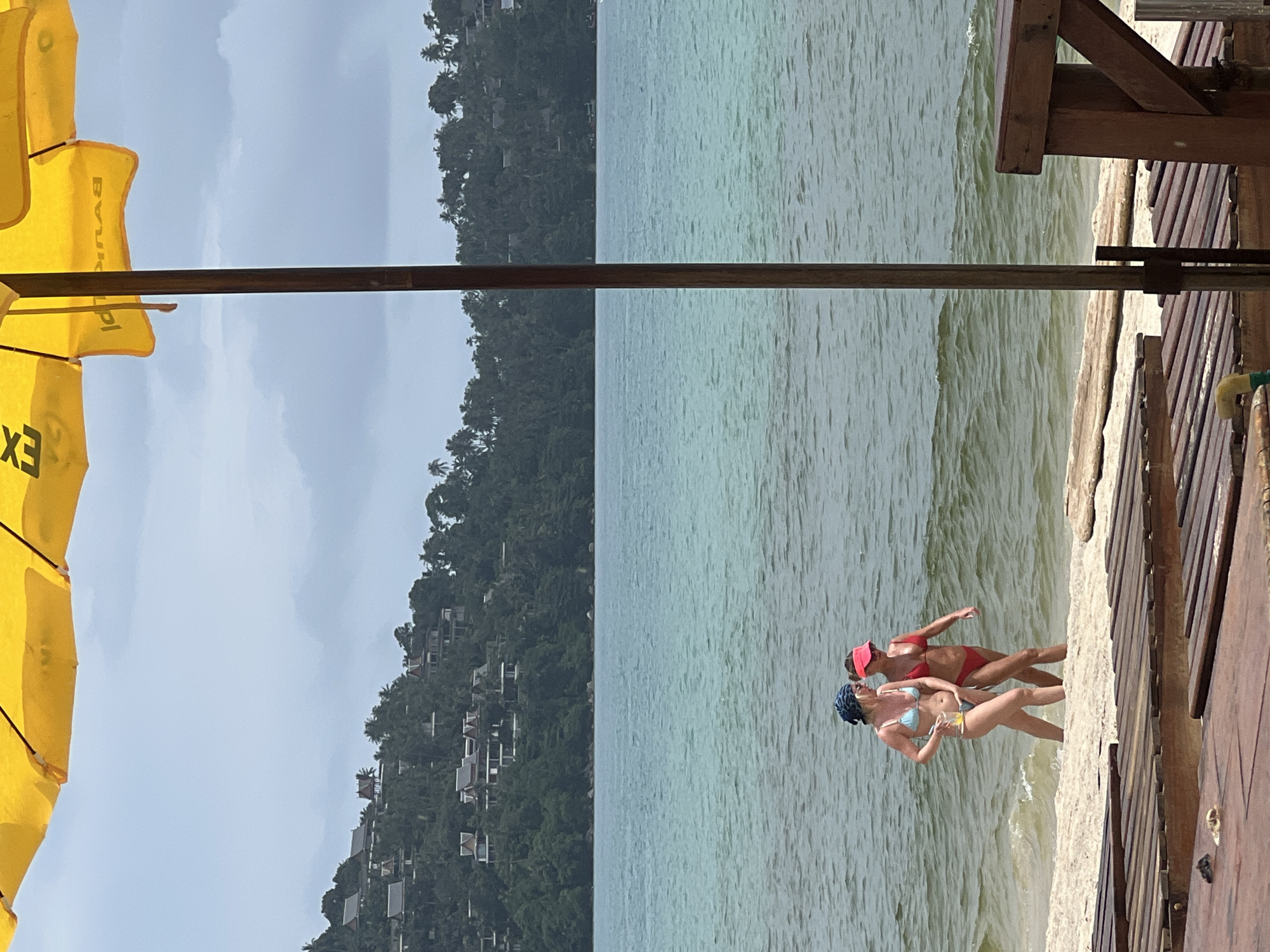 This image shows two women in swimsuits on a beach. The woman in the foreground is wearing a light blue bikini and a dark blue bandana. She is holding a drink with a straw. The woman behind her is wearing a red bikini and a pink visor. They are standing at the edge of the water, with the ocean in front of them and a densely forested hillside with buildings in the background. To the right of the women is a wooden deck or pier. The scene appears to be a tropical beach. The lighting suggests it is daytime. There is a large yellow umbrella on the left side of the image, with some black lettering visible on it.