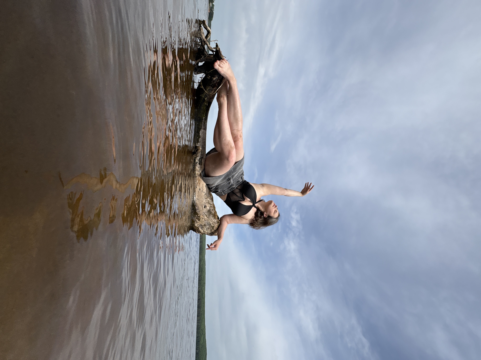 A woman is sitting on a tree trunk partially submerged in water. She is wearing a black top and gray shorts. Her legs are extended towards the left side of the image, while her arms are outstretched towards the right. The tree trunk appears to be positioned at the water's edge. The background consists of a cloudy sky. The water is brown. The location is in the city of Chaikovsky, Russia.