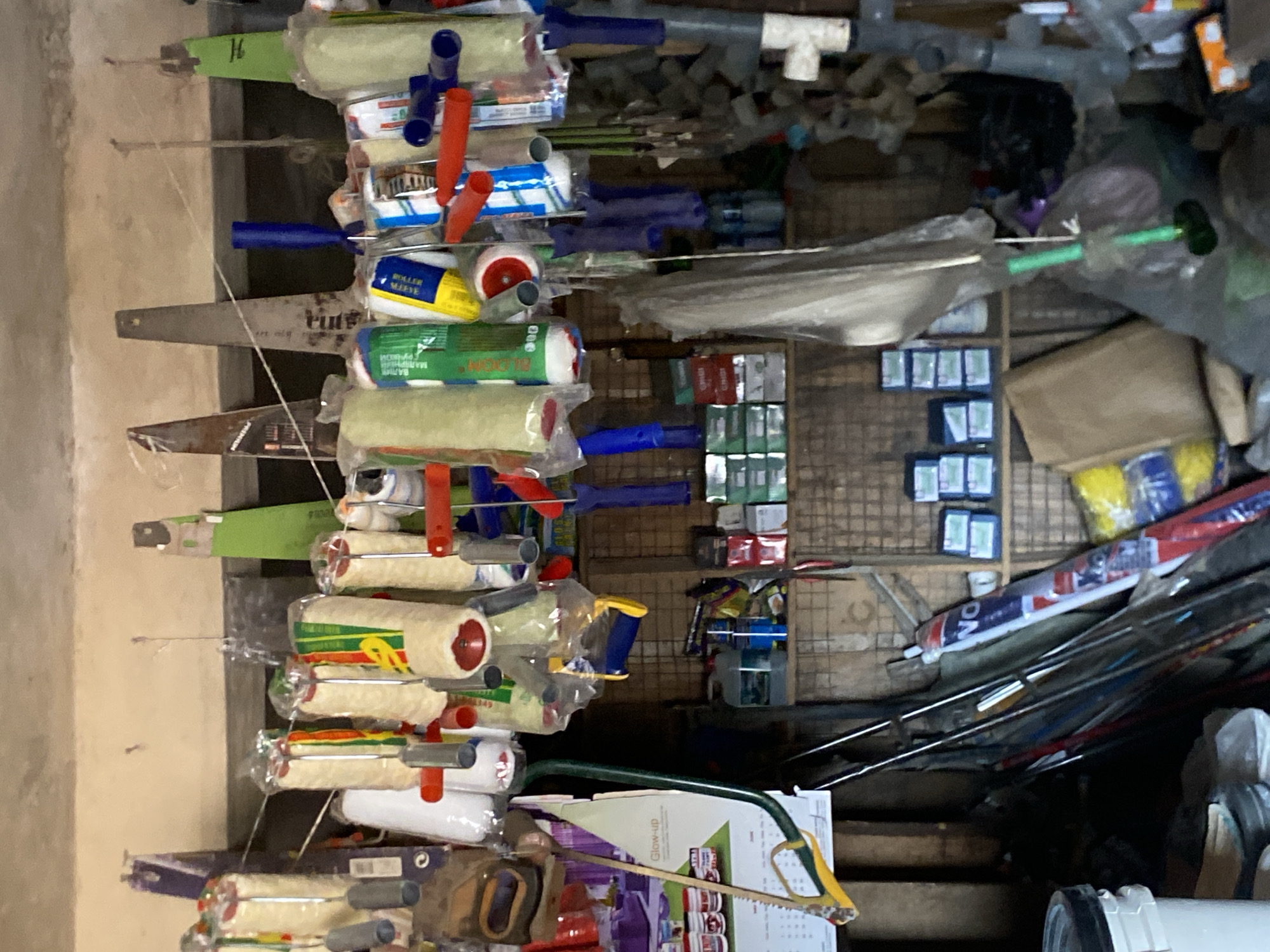 The image shows a cluttered storage or retail space in Bukuru, Nigeria. The foreground displays rows of paint rollers hanging on metal rods, with a variety of colors and packaging. Behind the rollers, the scene transitions to shelves filled with assorted hardware and construction materials, including pipes, small boxes, and various tools. The overall environment appears to be a small shop or storage area.