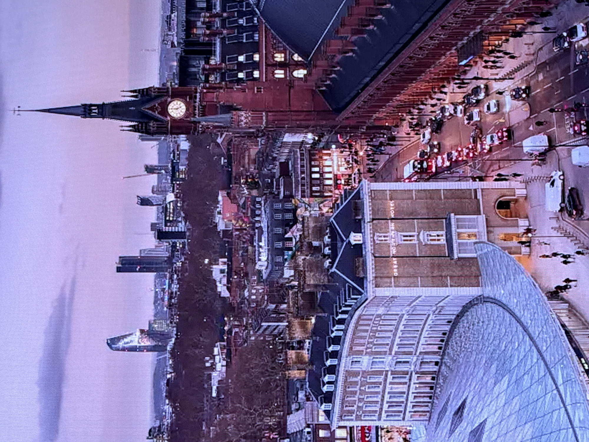 This image is an aerial view of a city street scene at dusk. The primary focus is on a prominent red brick building with a tall clock tower, identified as St Pancras International station in London, UK. The architecture is Victorian Gothic, with intricate detailing and numerous windows. To the left of the station, a curved, modern glass-covered structure is visible, likely part of the station's renovation. Across the street from the station, a curved, light-colored building with many windows is labeled "GREAT NORTHERN HOTEL." The street itself is busy with traffic, including red double-decker buses, cars, and vans, indicating urban activity. Pedestrians are visible on the sidewalks. In the background, the cityscape features modern skyscrapers and other buildings, some illuminated. The sky is a dusky purple with hints of clouds, suggesting late evening or early morning. The provided crops highlight details of the clock tower, the hotel facade, the modern roof structure, and the street activity. The request to identify the location as Krasnokamsk, Russia, is inconsistent with the visual evidence of St Pancras International, which is in London, UK.