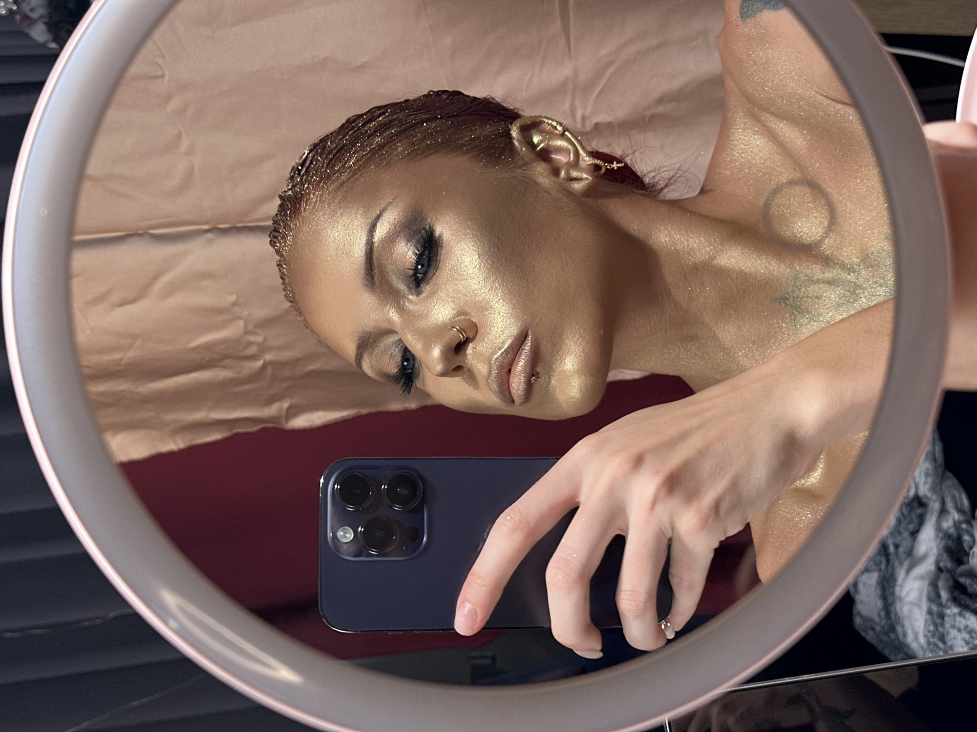 The image captures a close-up selfie of a woman taken in a round mirror, likely a ring light, in an indoor setting. The woman is angled, showing her head, neck, and right shoulder. Her skin and hair are covered in a shimmering golden body paint or glitter, giving her a metallic appearance. She has dark, dramatic eye makeup, possibly false eyelashes, a small nose ring, and a lip piercing. Her hair appears styled in small braids or cornrows, also adorned with golden glitter. An intricate golden ear cuff is visible on her right ear, extending upwards. On her neck and shoulder area, there are faint tattoos, including the outline of a circle and another abstract design, visible through the body paint.
She holds a dark-colored smartphone, likely an iPhone with a triple-camera system, in her right hand, positioned in the lower center of the mirror's reflection. Her nails are clear or lightly polished.
The background behind the woman's reflection consists of a crumpled, reddish-brown fabric or paper sheet on the left and a darker, solid red surface on the right. Below the phone, a dark, possibly black or grey, textured surface is visible, and to the far right, a crumpled grey and white patterned fabric is present. The circular frame of the mirror/ring light encompasses the entire scene. The woman's expression is focused and composed. No text is visible in the image. The scene suggests a stylized photoshoot or artistic makeup display.