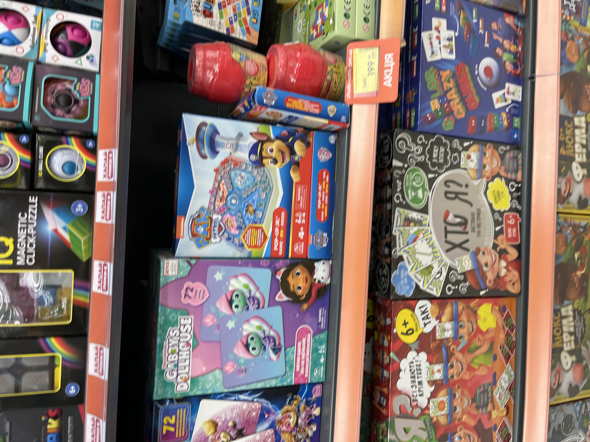 The image shows a retail shelf displaying a variety of children's toys and games. The products are predominantly board games and puzzles, with packaging featuring colorful cartoon characters and illustrations. Some of the visible brands include "Gabby's Dollhouse" and "Paw Patrol." There is a sale sign visible in the lower right portion of the image, displaying "199" and the word "АКЦІЯ" (AKTSYA), which translates to "Sale" or "Promotion" in Ukrainian. This suggests the location is a store in Ukraine. The overall environment appears to be a toy aisle within a retail establishment. There are no people clearly visible in the image. The lighting is bright, typical of a retail setting. The arrangement of the products indicates they are organized by category on shelves. The presence of Ukrainian text on the sale sign strongly suggests this scene is located in Kharkiv, Ukraine.