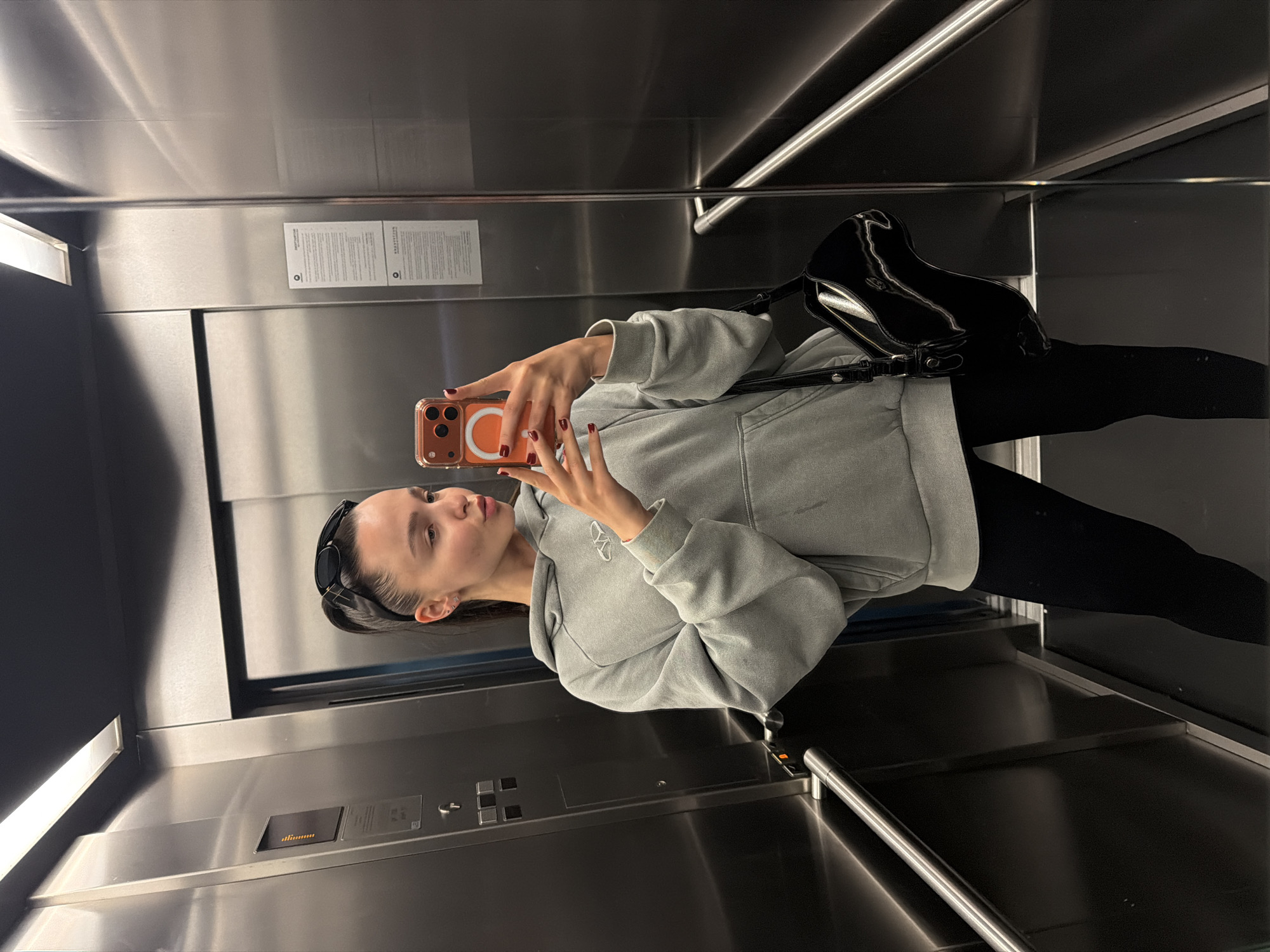 A young woman is captured in a mirror selfie inside an elevator, likely located in Sofia, Bulgaria. She is facing the reflective metal wall, holding an orange-cased smartphone with a white circular MagSafe accessory to take the picture. Her fingernails are painted red. She has dark hair pulled back, with dark sunglasses resting on top of her head.
The woman is wearing a light grey, possibly faded, hooded sweatshirt (hoodie) with a white, stylized 'W' or abstract logo on the left chest. She pairs this with dark leggings or pants. A shiny black shoulder bag is visible, hanging from her left shoulder.
The elevator interior features brushed metallic walls and a metallic door. Bright linear lights illuminate the space from the upper and lower edges. On the right wall of the elevator, a control panel is visible, equipped with buttons and a digital indicator displaying a series of horizontal lines. A metallic handrail runs horizontally across the wall beneath the control panel. Two white papers with dense blocks of text, resembling informational or safety instructions, are affixed to the elevator wall above the control panel. The overall setting is modern and functional, characteristic of a building elevator. The woman's expression is neutral, focusing on her phone screen to compose the selfie.