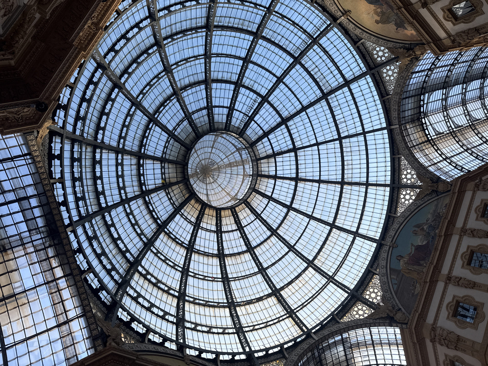 The image depicts the glass dome of the Galleria Vittorio Emanuele II in Milan, Italy, viewed from below. The dome is constructed of metal framing and glass panels arranged in concentric circles, converging at a central oculus. The architectural details of the surrounding building, including ornate cornices and painted frescoes, are visible at the edges of the frame. The sky is visible through the glass. There are no people visible in the frame.