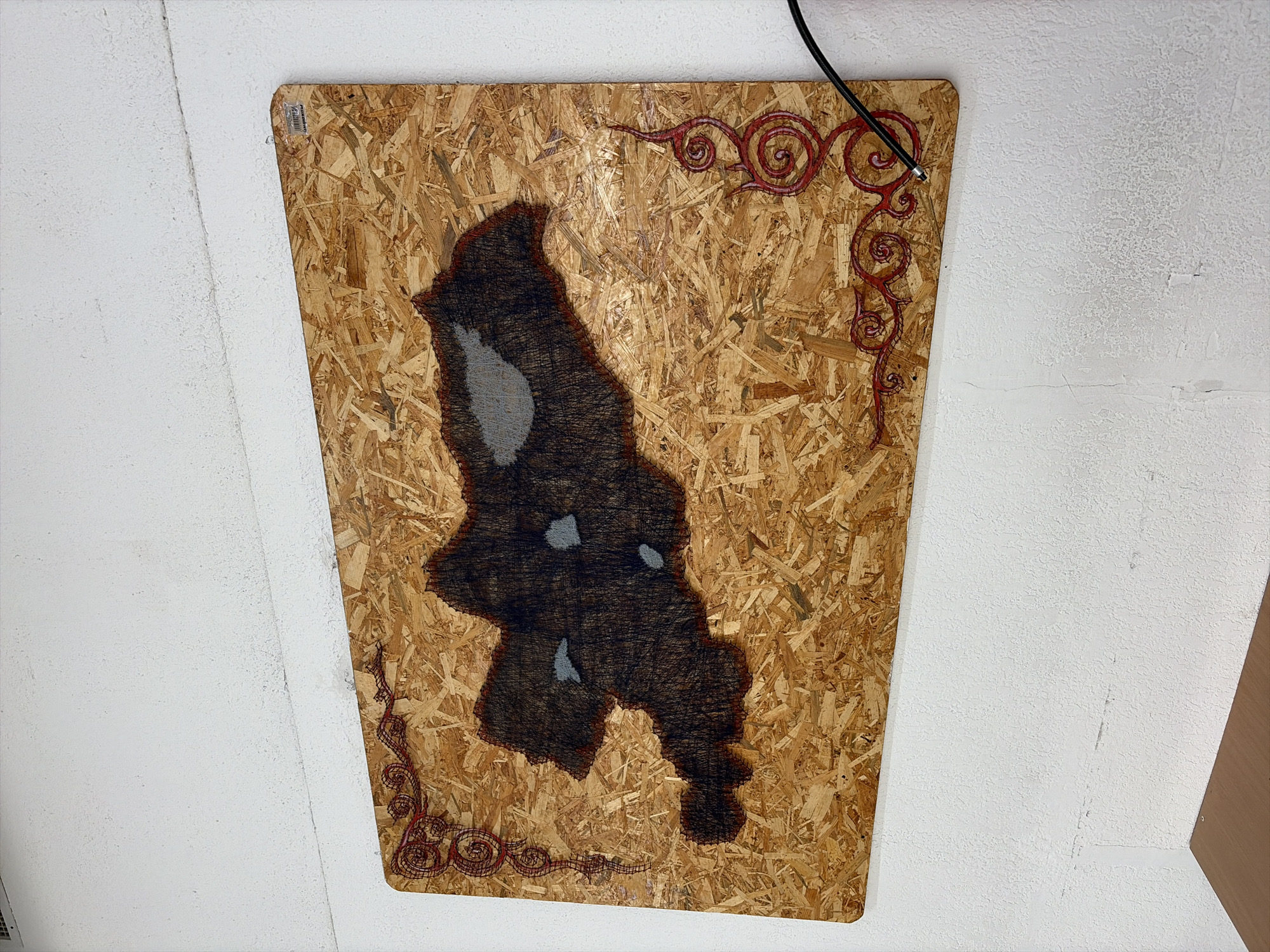 The image shows a piece of art on a textured white wall, likely indoors. The artwork is mounted on a rectangular piece of oriented strand board (OSB). The OSB has a light brown, wood-chip texture. The artwork itself is a silhouette filled with dark thread, resembling a dark, irregular shape with some lighter gray splotches within it, potentially representing bodies of water or shadows. Red, swirling decorative patterns are present along the left and right edges of the OSB board, framing the main dark silhouette. A black cable extends from the lower right corner of the OSB board and disappears out of frame to the right. The wall has some subtle texture and appears to be a pale, off-white color. There is a dark wooden surface visible at the bottom of the frame, suggesting a table or shelf. The overall impression is of an art installation or display within a room. No specific location in Bishkek, Kyrgyzstan can be identified from the visual information alone.