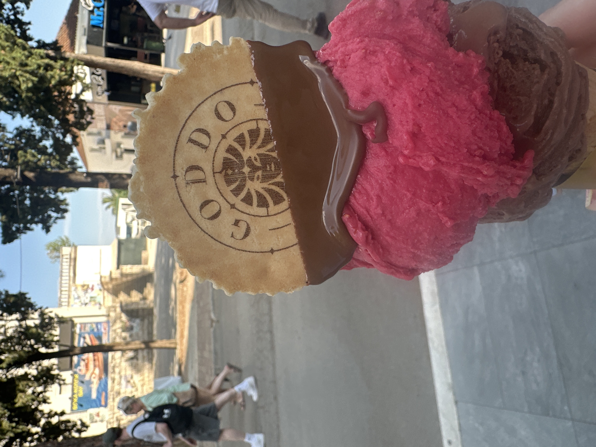 A partially rotated eye-level shot of a two-scoop gelato cone, featuring a circular wafer with chocolate drizzle, held in a hand, and viewed against the backdrop of a busy street scene in Budva, Montenegro. The top scoop is vibrant pink, the bottom is a deep chocolate color. The wafer, bearing the letters "GODO", is partially covered with dark brown chocolate drizzle that extends onto the pink ice cream.
In the background, pedestrians walk on a grey sidewalk and a street with various shops and buildings. A blue sign reading "Mr. C" is visible above one of the storefronts. Trees with green foliage line the street, contributing to the urban landscape. Further down the street, buildings with light-colored walls and architectural details can be seen under a bright sky.