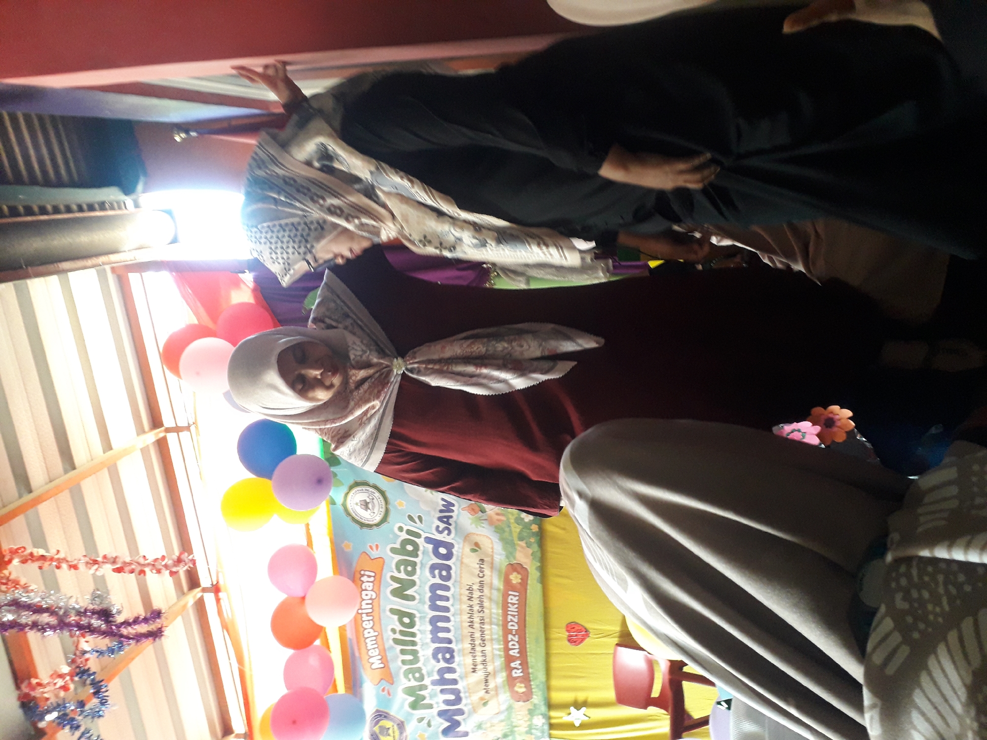 In the city of Cankuang, Indonesia, a photograph captures a celebration likely related to Maulid Nabi Muhammad SAW. Two women, both wearing hijabs, stand prominently. The woman on the left is dressed in a maroon robe and a patterned scarf. The woman on the right wears a black garment and a patterned scarf. Behind them, a banner reads "Memperingati Maulid Nabi Muhammad SAW" and further text, "Meneladani Akhlak Nabi, Mewujudkan Generasi Saleh dan Ceria" and "RA ADZ-DZIKRI." The background is adorned with colorful balloons and festive decorations, suggesting a lively event. In the foreground, a person is partially obscured by draped fabric, and a red chair is visible. The lighting is bright, with some areas appearing overexposed.
