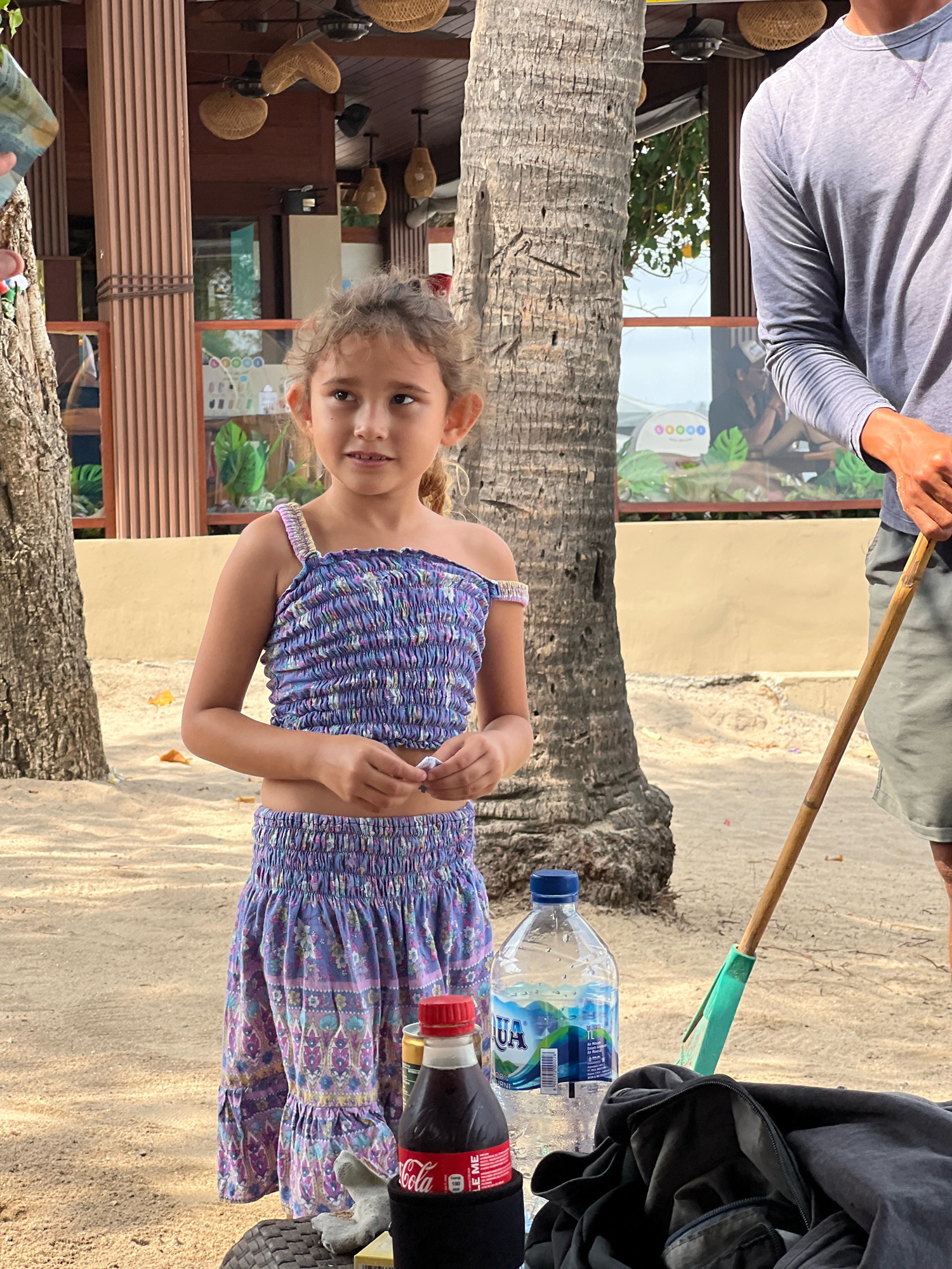 In Kuta, Indonesia, on a sandy beach, a young girl with curly hair stands next to a palm tree. She is wearing a purple floral smocked top and a matching skirt. Her hands are clasped in front of her. To her right, a person in a grey long-sleeved shirt and green shorts stands holding a bamboo pole with a green attachment. In the foreground, a bottle of Aqua water and a bottle of Coca-Cola are on a table, along with a black bag. Behind the subjects, a building with wooden pillars and woven ceiling decorations is visible. The environment suggests an outdoor dining or recreational area.