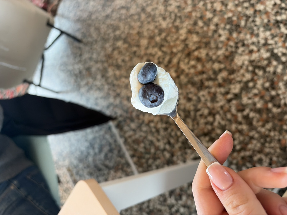 The image displays a hand holding a stainless steel spoon, oriented diagonally from the lower left towards the upper right. The spoon's bowl contains a white, creamy substance topped with two dark blue blueberries. The hand exhibits manicured fingernails with a light pink base and white tips. The background is out of focus, indicating an indoor setting. To the upper left, a light-colored table edge is partially visible. Centered in the mid-ground, a person's lower torso and legs are obscured but visible, dressed in blue denim jeans and dark fabric, positioned near the dark metal frame of a chair. The floor is a speckled, multi-tonal surface, likely terrazzo. This scene takes place indoors in La Clusaz, France.