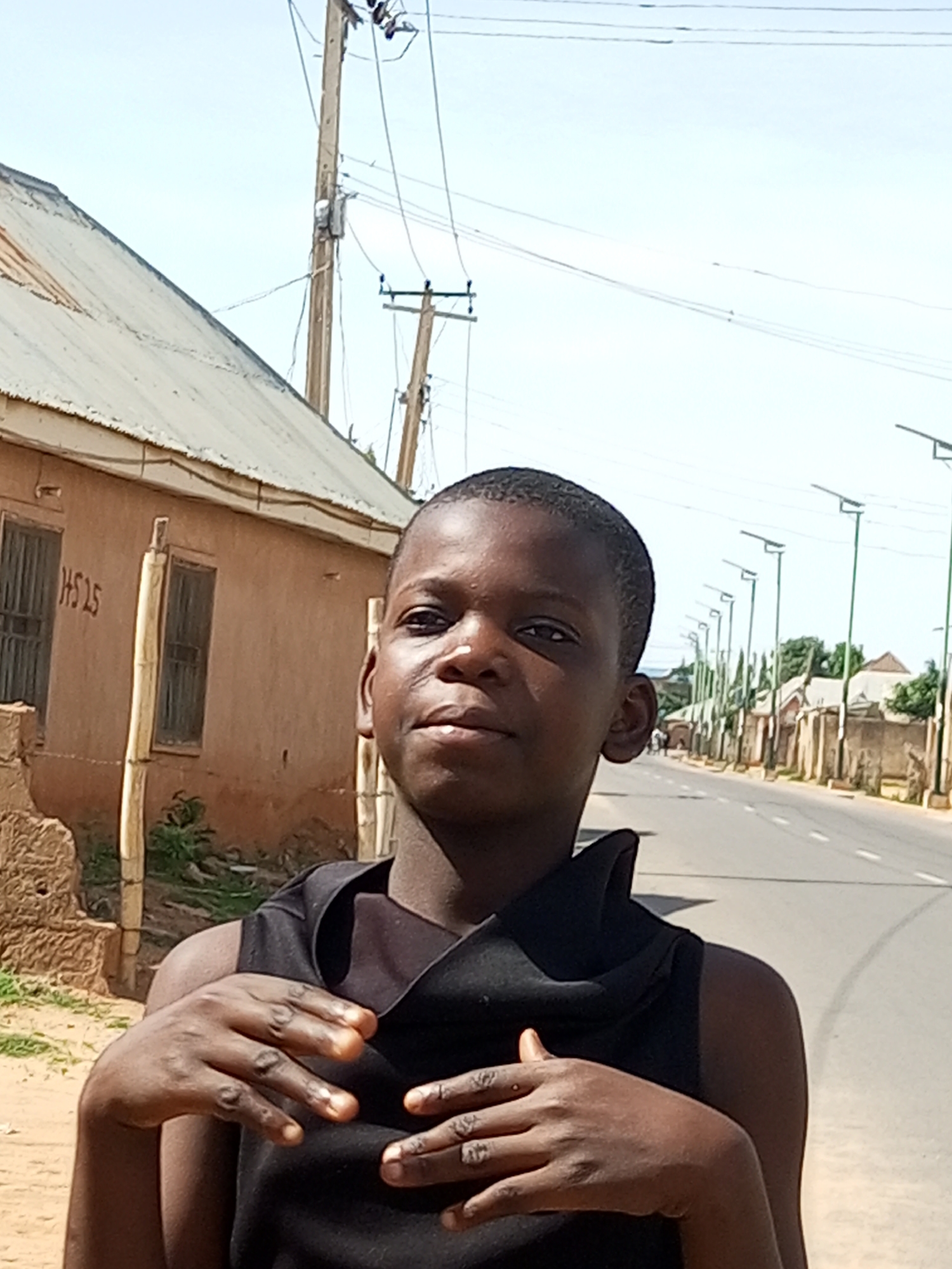 A young boy stands in the foreground, facing the camera with his arms bent and hands clasped in front of his chest. He is wearing a black sleeveless top. His head is tilted slightly to his left, and he has dark skin and short, dark hair. Behind him, a dirt path runs alongside a paved road lined with streetlights. To the left of the path is a low-rise building with a corrugated metal roof and mud-colored walls. The building has two windows with metal bars. A wooden pole with wires is visible near the building. The sky is clear and bright. The scene appears to be in a residential area of Tempure, Nigeria.