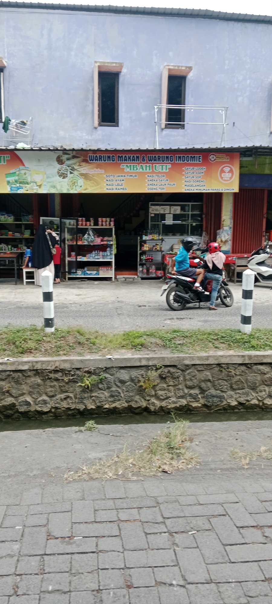 The image captures a street scene in Maguwoharjo, Indonesia, with a focus on a food and noodle warung named "MBAH UTI". The warung is a single-story building with a light blue facade and two tall, narrow windows on the upper level. A banner above the warung entrance advertises "WARUNG MAKAN & WARUNG INDOMIE" and lists various menu items such as "SOTO JAWA TIMUR", "NASI AYAM", "NASI LELE", "NASI RAMES", "NASI TELOR", "ANEKA SAYUR", "ANEKA LAUK PAUK", "OSENG TERI", "SAYUR LODEM", "SAYUR ASEM", "SAYUR SOP", "MAGELANGAN", and "ANEKA MINUMAN PANAS & DINGIN". The warung has shelves stocked with goods, including bottled drinks and packaged food items.
In the foreground, a black motorcycle is parked on the side of the road, with two individuals on it. One person, wearing a blue shirt and a black helmet, is seated on the back, while another, wearing a pink headscarf and jeans, is on the front, seemingly preparing to operate the motorcycle. Another person, wearing a black hijab and a light-colored dress, is standing near the entrance of the warung, looking towards the interior. A younger child in a blue shirt is standing near a table to the left of the warung entrance.
The environment includes a paved road, a low concrete wall with a stone base bordering a narrow, murky body of water, and patches of grass and weeds. Two black and white striped poles are positioned on the side of the road, one in the foreground and another further down. The overall impression is a bustling local eatery with customers arriving and departing on motorcycles.