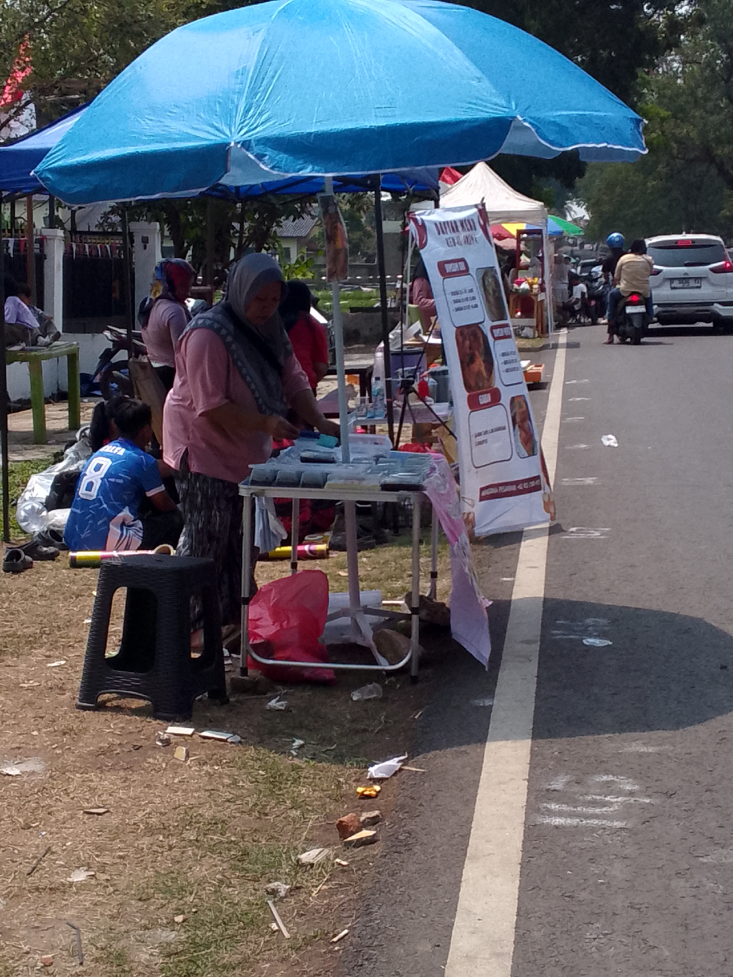 The image depicts an outdoor scene in Mandalawangi, Indonesia, likely a street market or roadside stall area. A large blue umbrella shades a vendor and her display. The vendor, wearing a pink shirt and a headscarf, is interacting with items on a table. Beside her, a young person in a blue and white jersey with the number "8" is sitting on the ground. Various items, including bags and what appear to be snacks or food items, are spread around the area. In the background, there are other stalls, people, and a white car driving on the road. A white banner with Indonesian text and images is prominently displayed, advertising food. The ground is a mix of dirt and grass, with some debris scattered around. The overall impression is of a busy, informal commercial setting.