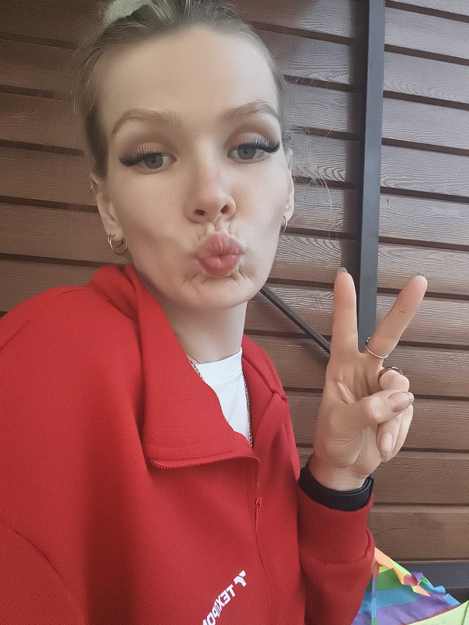 A young woman is photographed from the chest up, facing the camera with a pout and her right hand raised in a peace sign. She has her hair pulled up in a bun and is wearing makeup, including false eyelashes. Her attire consists of a red zip-up hoodie over a white t-shirt. The background is a wooden wall, suggesting an outdoor setting. There is no indication of the location being in Русский Вожой, Russia.