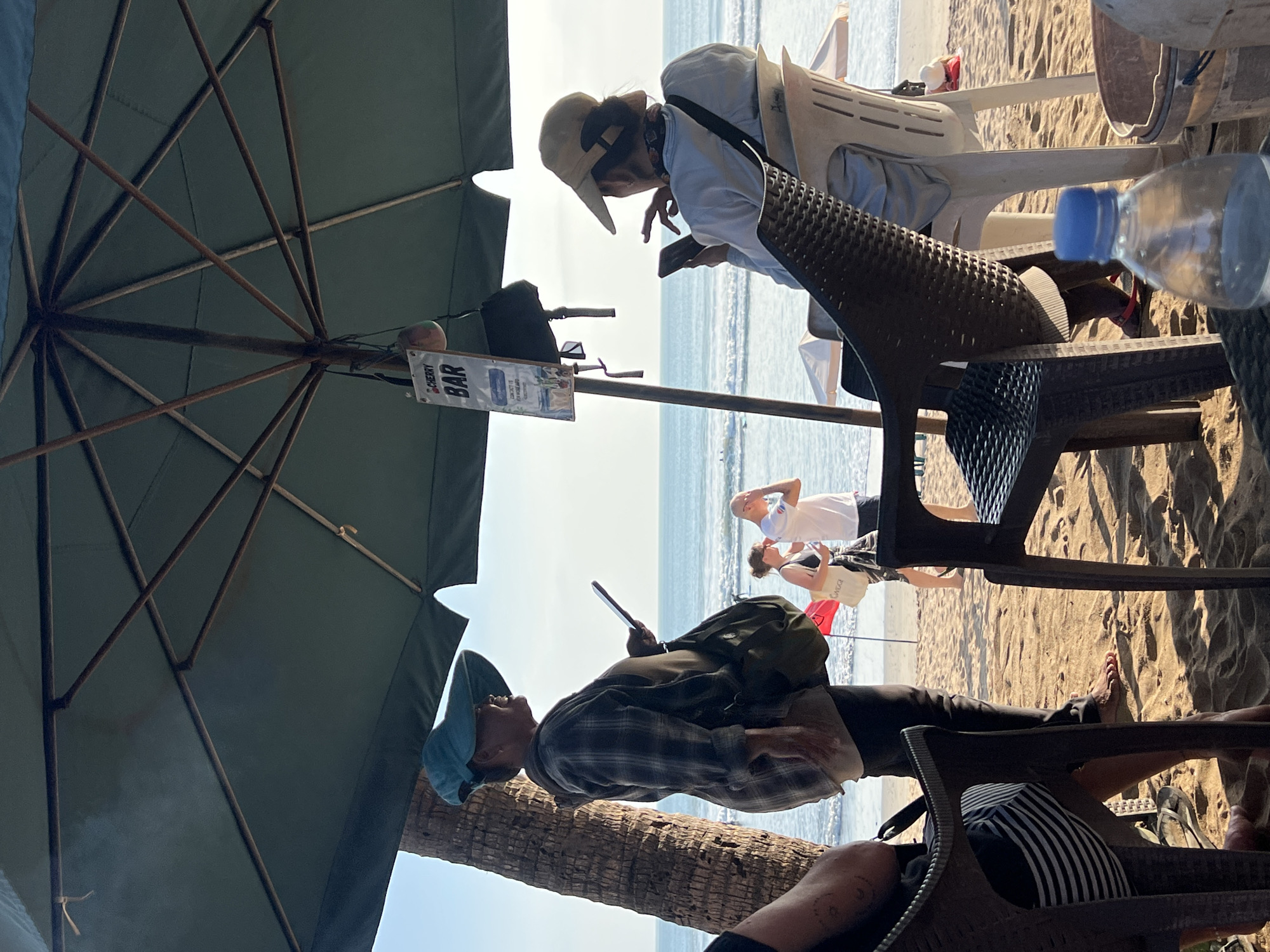 The image depicts a beach scene in Kuta, Indonesia, likely near a bar under a large green umbrella. In the foreground, the legs of a person wearing a striped shirt and flip-flops are visible, seated in a dark plastic chair. Another dark plastic chair and a white plastic chair are also present. On a table in the foreground, a plastic water bottle with a blue cap is prominent.
A woman wearing a wide-brimmed blue hat and a plaid shirt is standing and looking to her right, holding a tablet or phone. She has a green bag slung over her shoulder. Behind her, a man in a white t-shirt and a woman in a black tank top are walking on the beach. Further back, a person in a beige hat is seated, looking at their phone. The ocean and waves are visible in the background, under a clear sky. A sign hanging from the umbrella reads "CHERRY BAR." A coconut and a black bag are also hanging from the umbrella. Palm trees are visible on the left side of the frame. The sand on the beach is light brown and textured, with shadows cast by the umbrella.