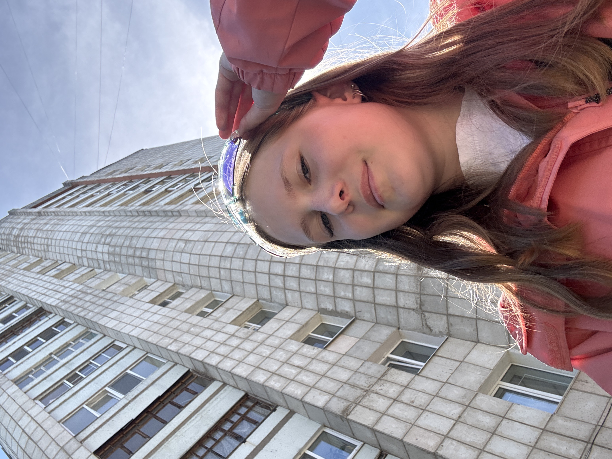 A young person is taking a selfie from a low angle, with a tall building in the background. The building is characterized by a gray tiled exterior and numerous windows arranged in horizontal rows. The individual is wearing a pink jacket and has long brown hair. They are touching their hair near their forehead with their hand. The sky is visible in the upper portion of the frame. The location is Chaikovsky, Russia based on previous information.