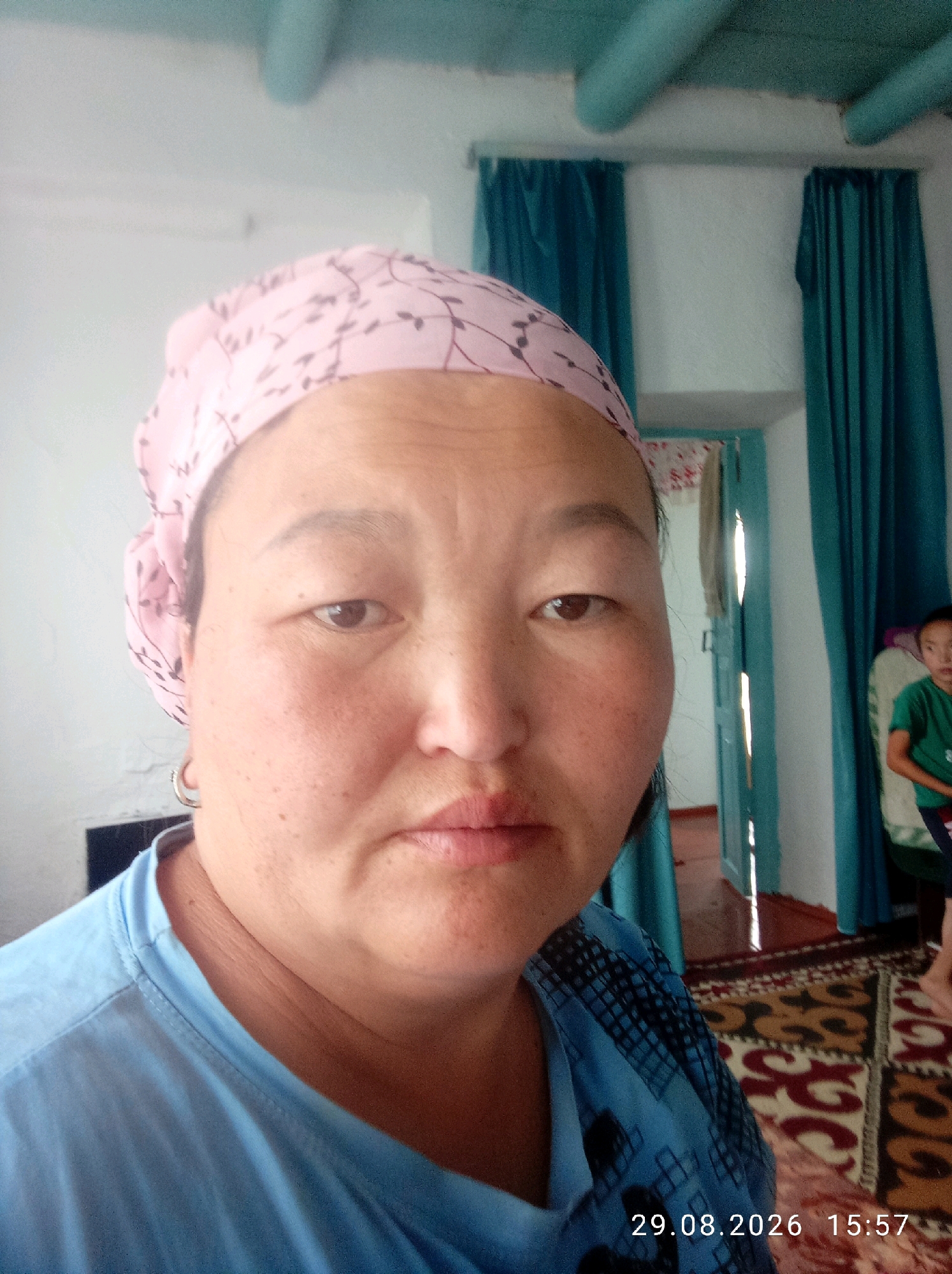 A middle-aged woman wearing a light pink headscarf with a dark leaf pattern and a blue t-shirt is photographed from the chest up, facing the camera. She has fair skin with freckles and wrinkles, dark eyes, and a pinkish-toned mouth. A silver hoop earring is visible on her left ear. In the background, teal curtains hang on either side of an open doorway. A child in a green t-shirt is seated on a chair to the right. The room has a white ceiling with exposed light blue beams and a patterned rug on the floor. The date stamp "29.08.2026 15:57" is visible in the bottom right corner.