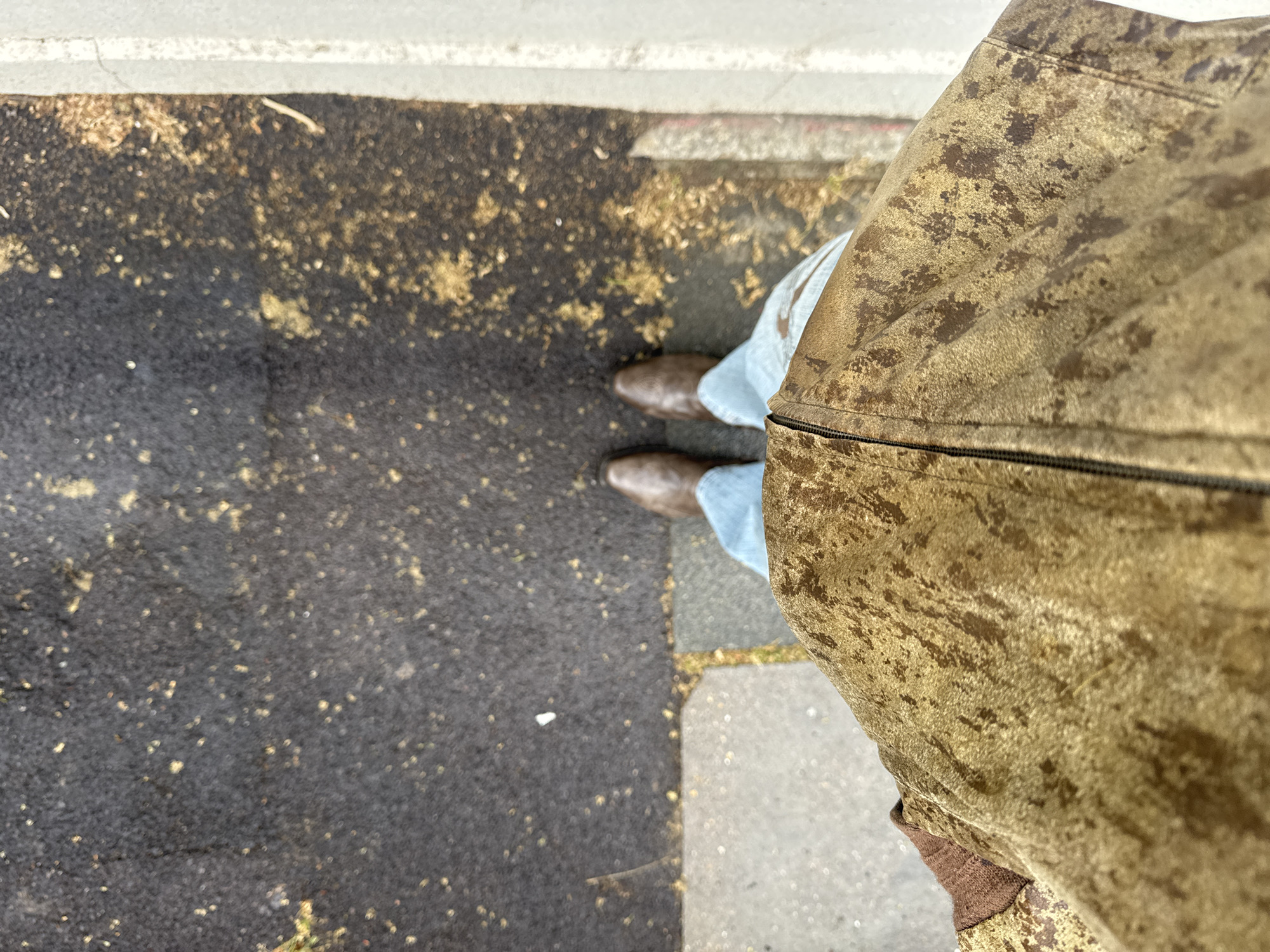 A high-angle shot depicts a person standing on a sidewalk, likely in London, United Kingdom, as the street surfacing is commonly found in London. The individual wears light blue jeans and brown leather boots. The right side of the frame is dominated by a textured, tan-colored jacket. The sidewalk transitions from dark asphalt, covered with yellowish debris, to concrete slabs closer to the person. A gray curb borders the asphalt.