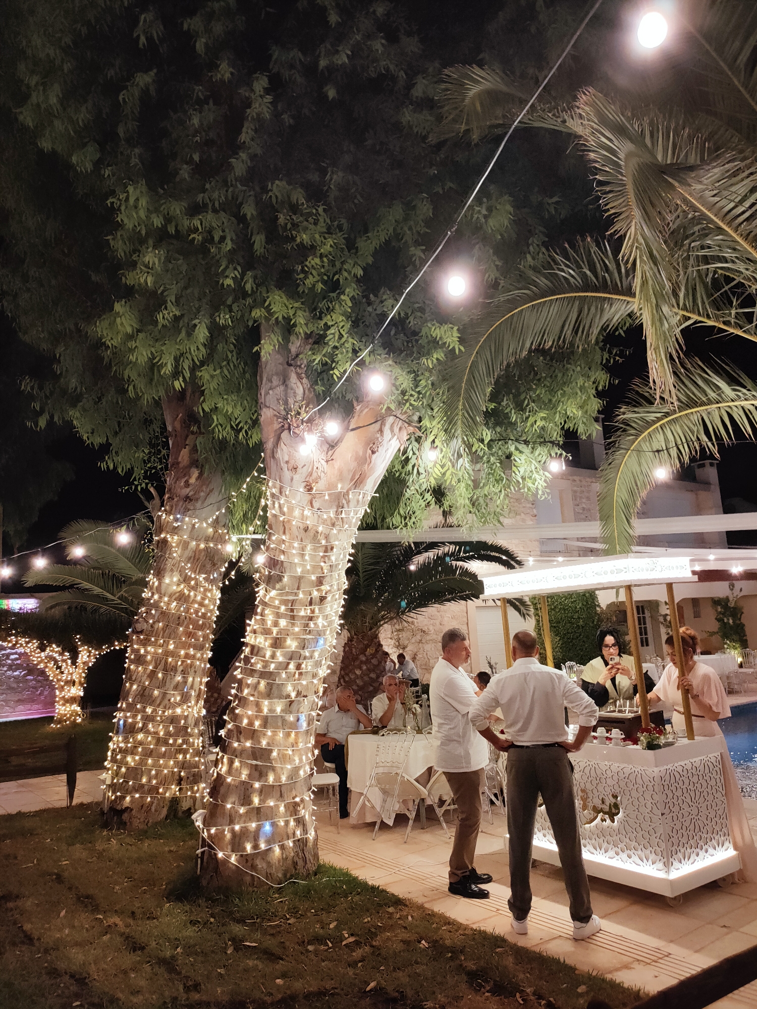 This is a nighttime outdoor scene in Cebalat, Tunisia, likely at a reception or party. The foreground features a grassy area with two large tree trunks wrapped in strings of warm white fairy lights. Beyond the trees, a white, ornate bar or counter is set up, attended by several people. A woman in a light pink dress is behind the bar, and a woman in a yellow and black outfit is holding a phone and looking at it. Two men in white shirts stand near the bar, facing away from the camera. In the background, there are tables set up with chairs, suggesting a dining or seating area. Palm trees and other foliage are visible, along with strings of lights illuminating the space. The overall ambiance is festive and illuminated by artificial lights.