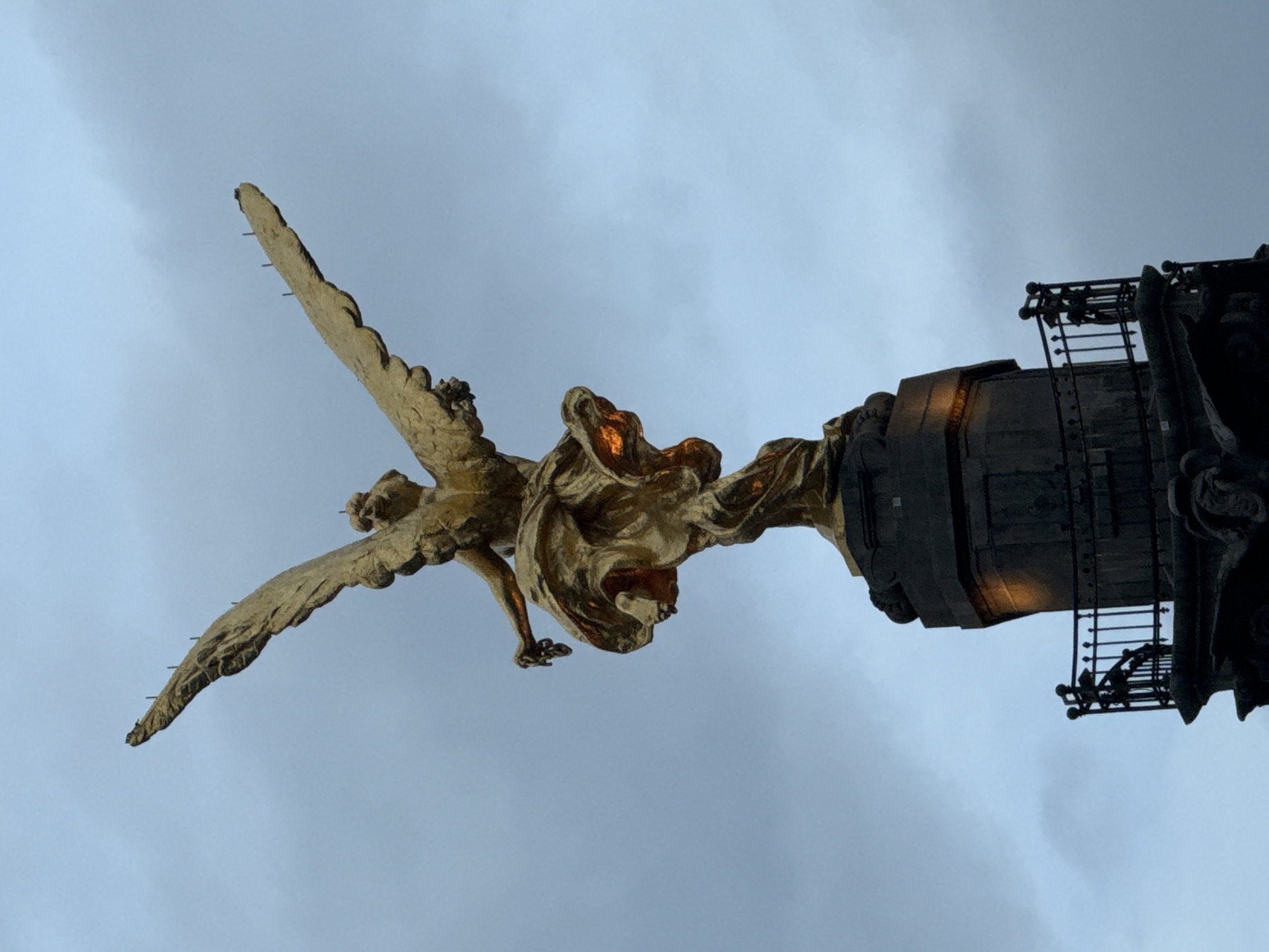 The image depicts the iconic golden statue of the Angel of Independence atop its column in Mexico City, Mexico. The statue is viewed from below against a cloudy sky. The angel is depicted with outstretched wings. The column base is visible, including elements of its ornate structure and access ladder.