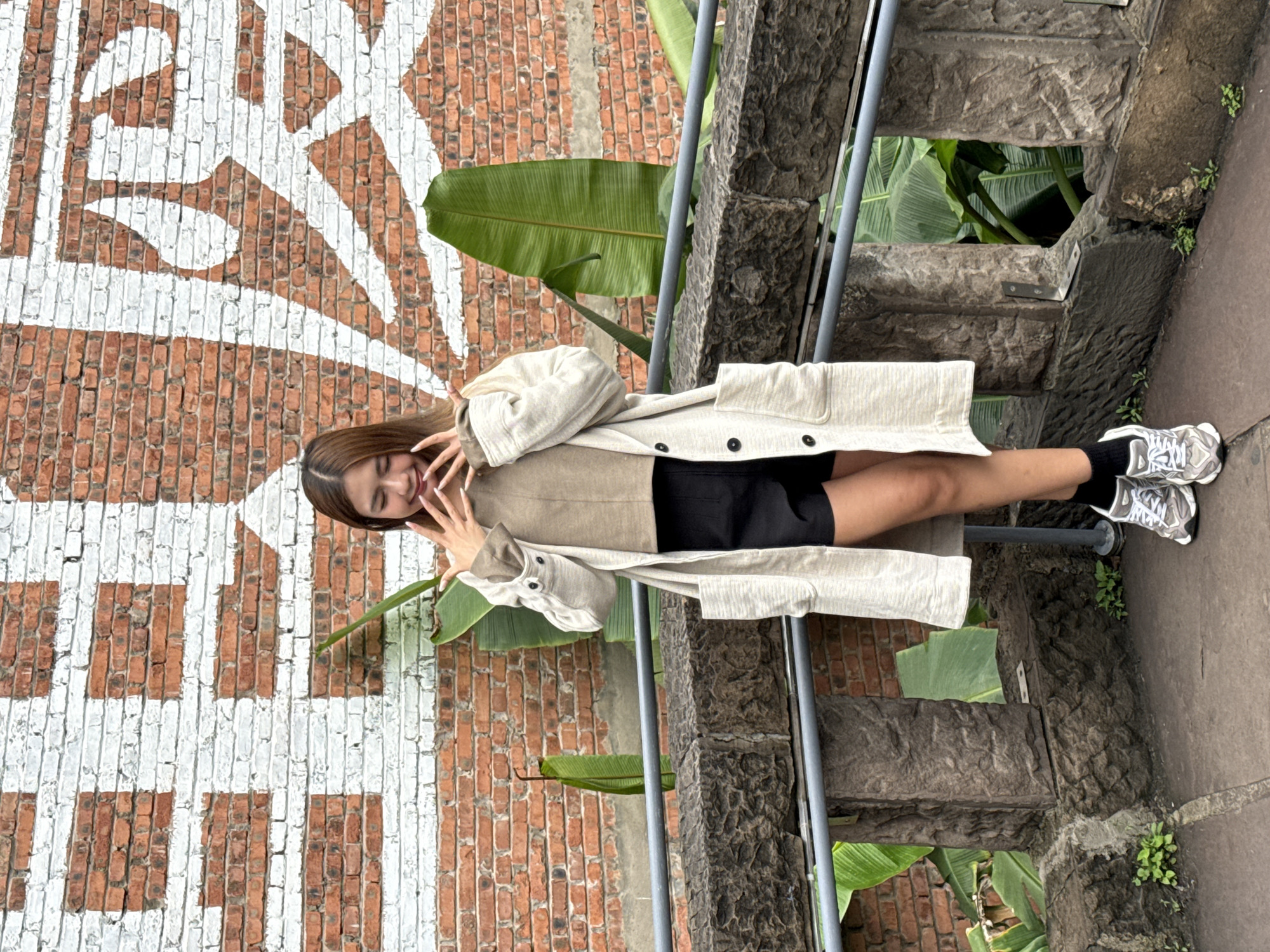 In Nan'an District, China, a young adult female stands facing the camera in an outdoor setting. She has light brown, straight hair and displays a closed-eye smile, with both hands raised to her face in a splayed finger gesture. She is dressed in a light beige long-sleeved overcoat with large front pockets and dark buttons, worn over a tan high-neck top, black shorts, black ankle socks, and grey and white athletic shoes. The background consists of a red brick wall partially painted with a large, stylized white geometric graffiti design. Large green tropical leaves are visible both behind her and interspersed with the brickwork. In the foreground, a dark grey, rough-hewn stone balustrade with two horizontal metal railings separates the subject from the immediate foreground. Small green plants are observed growing from cracks in the stone structure and on the dark paved ground.