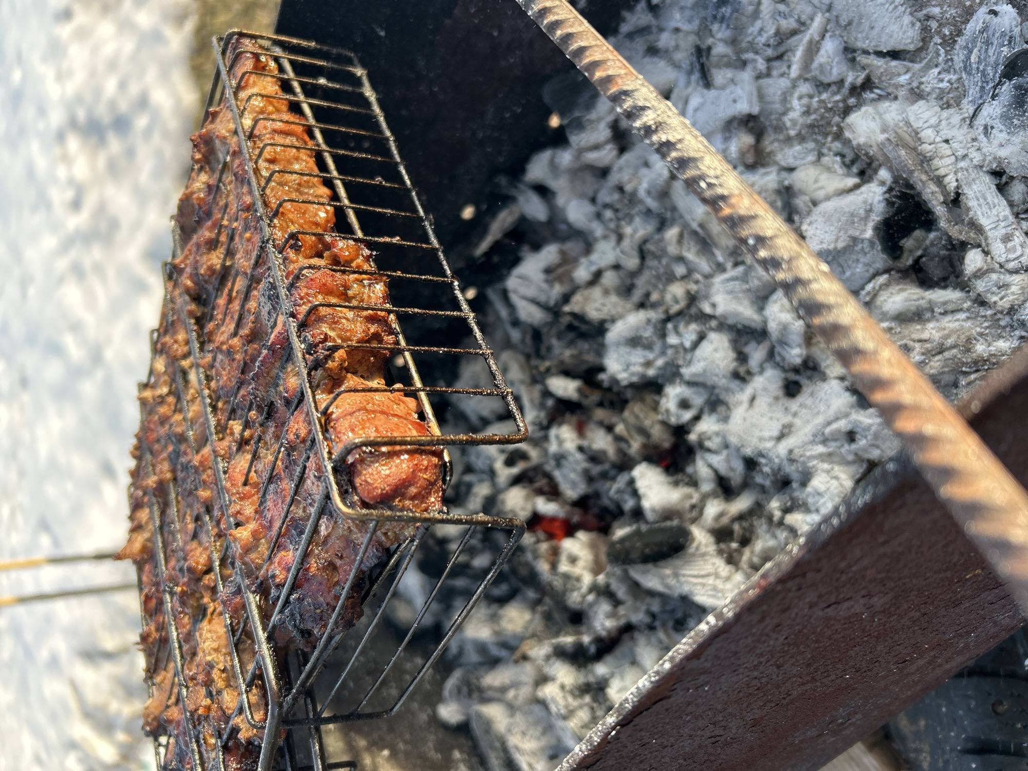 The image displays a close-up perspective of meat being grilled outdoors in the city of Noviy, Russia. A rectangular wire metal grilling basket, containing numerous pieces of reddish-brown meat with visible charring and some glistening surfaces, is positioned directly over a bed of glowing charcoal embers and grey ash. The charcoal is contained within a dark, utilitarian metal brazier or grill, with its upper edge showing signs of wear and rust. A rusty, ribbed metal rod is partially visible diagonally across the lower right foreground. The background is out of focus, revealing a snow-covered ground surface, suggesting a cold weather environment. Two slender, pole-like objects are indistinctly visible vertically in the distant background against the snow. No individuals are present in the scene.