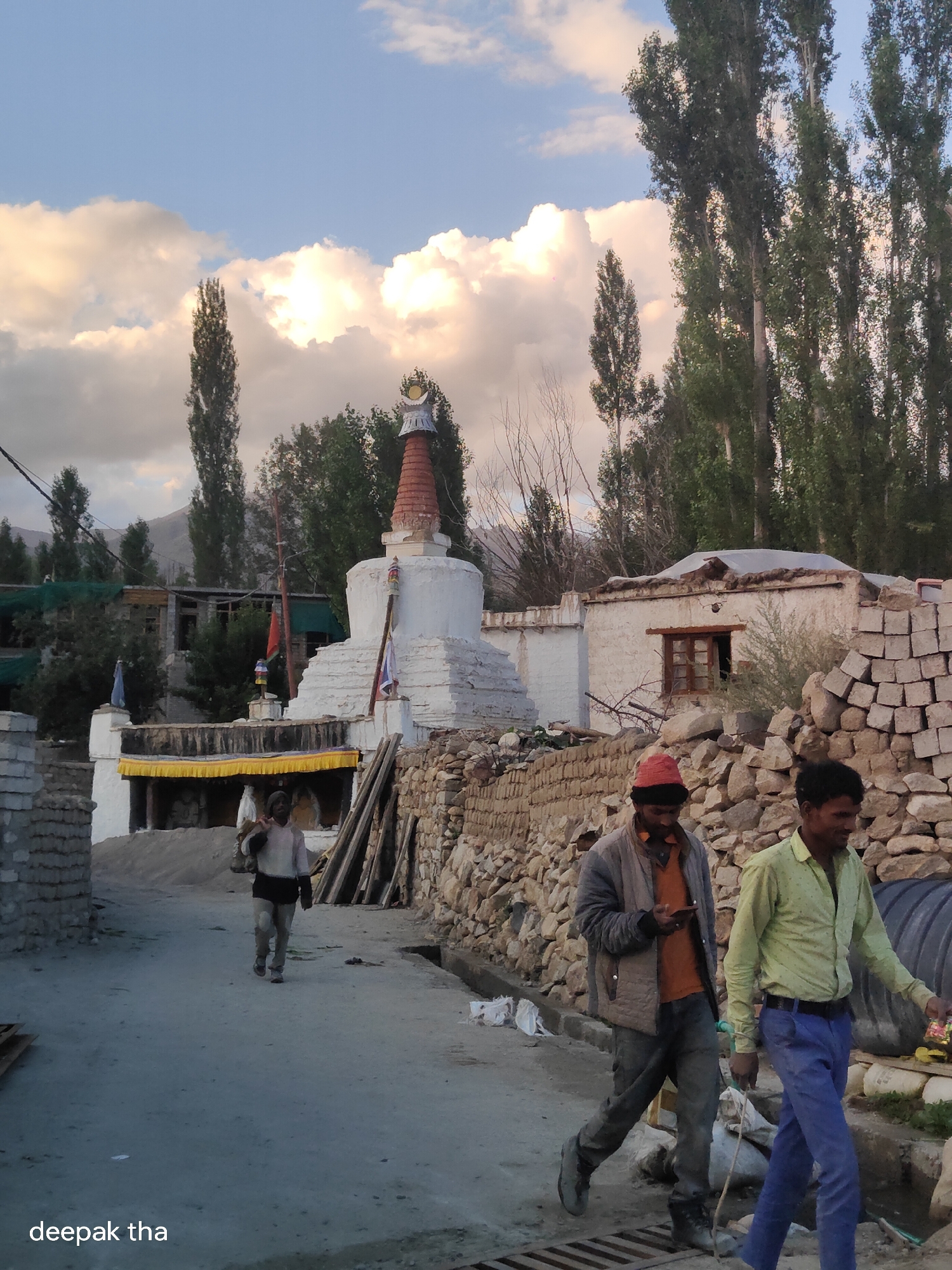 The image depicts a street scene in Leh, India, near a Buddhist stupa. The stupa is white and conical with red accents at the top, and flags are visible on its structure. A group of people are walking in the foreground. Two men are walking from right to left, one wearing a green shirt and blue pants, the other in a grey jacket over an orange shirt and jeans, holding a phone. Further in the background, a person is walking away from the camera carrying a large bundle on their shoulder. The street is paved and appears to be dusty. Buildings with white walls and traditional architecture line the street, some appearing to be under construction or repair with stacks of stones visible. Tall trees with green foliage are present in the background. The sky is a mix of blue with fluffy white and grey clouds, suggesting late afternoon or early evening light. The overall impression is of a somewhat rural or traditional area within a city.