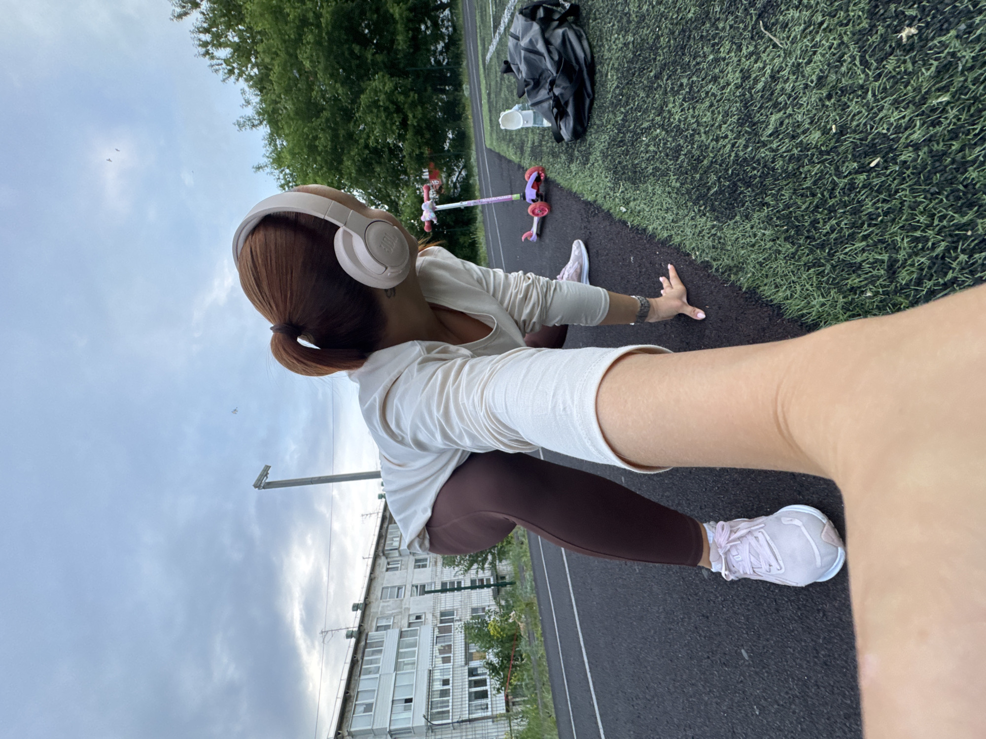 The image shows a person stretching on a track near a building in what appears to be the city of Chaikovsky, Russia. The person is wearing leggings, a light-colored shirt, headphones, and sneakers. A bag and a child's scooter are on the ground near the track. The sky is overcast. The background includes a building, trees, and a section of artificial turf. The image appears to have been taken with a wide-angle lens or from a close-up perspective.