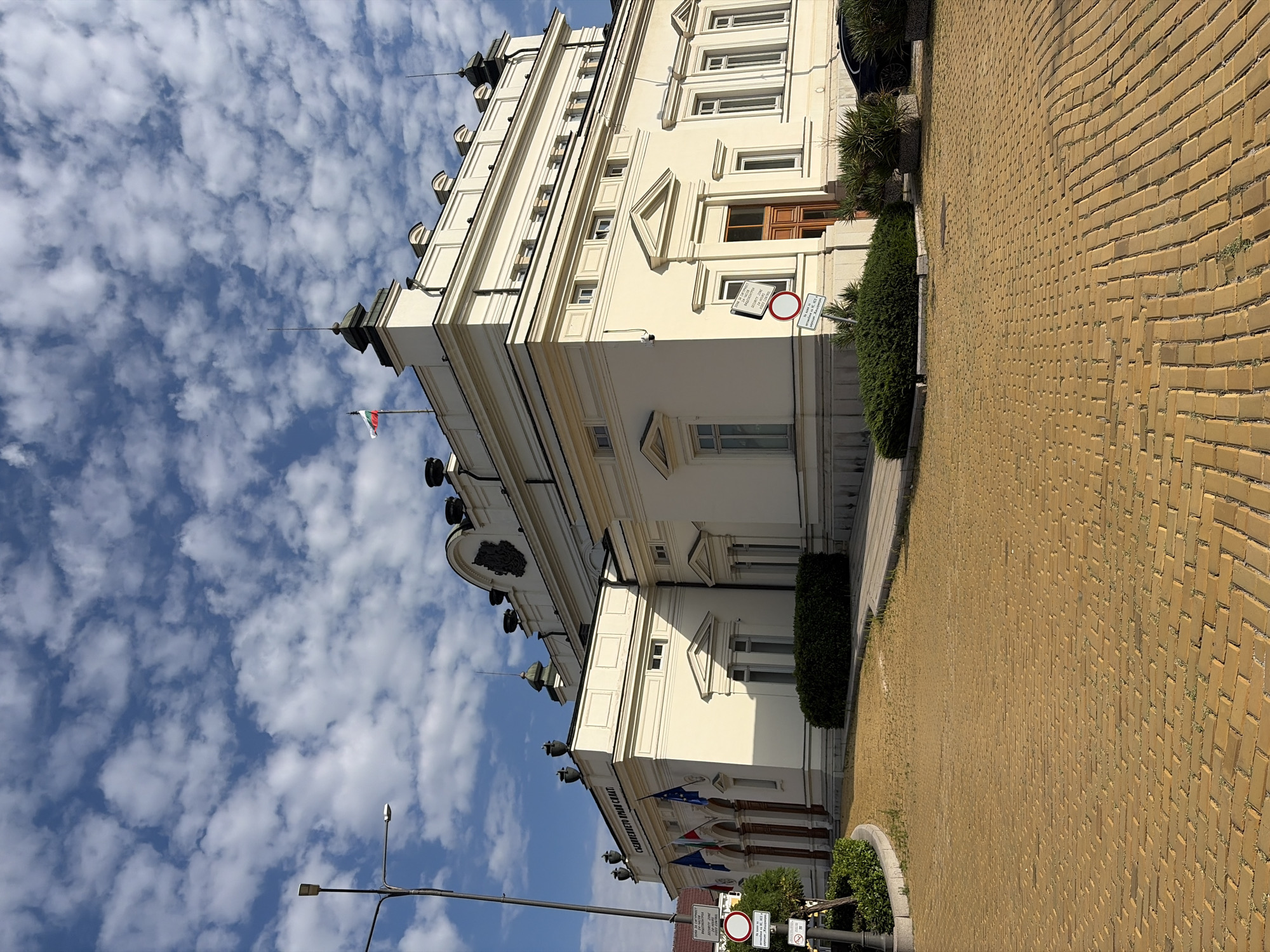 The image shows the National Assembly of Bulgaria, located in Sofia. The building is a large, pale yellow, neoclassical structure with an ornate facade. In front of the building, a cobblestone path made of yellow bricks leads to the entrance. Several flags are flying in front of the building, and there are signs and lampposts visible. The sky is blue with scattered white clouds. A black car is parked to the right of the building. Lush green bushes and trees are planted around the building.