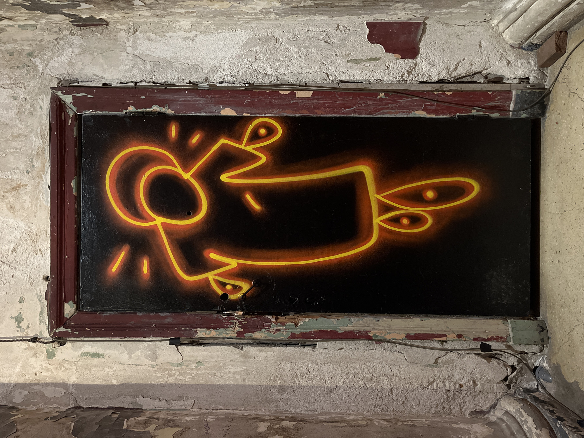 The image displays a deteriorated doorway in an interior setting within Saint Petersburg, Russia. A dark, vertically oriented door is centrally positioned, featuring a brightly illuminated, stylized anthropomorphic figure rendered in distinct orange and yellow outlines with radiating line elements around its head and limbs. The figure's head is circular, its body simplified, and two ovoid shapes are present at its lower section.
The door frame is heavily worn, with multiple layers of chipped paint, primarily dark red over lighter tones, exposing underlying wood and plaster. A small, brass-colored padlock is affixed to the left side of the door, approximately two-thirds up from the bottom.
The surrounding walls exhibit severe dilapidation, characterized by extensive peeling and flaking plaster and paint in shades of off-white, grey, and residual green and red. The exposed substratum is rough and textured. Faint Cyrillic text is faintly visible on the lower left wall. A thin black cable extends vertically along the left wall and continues along the floor. A grey, curved pipe or conduit is situated at the base of the left wall. A reddish brick rests on the floor adjacent to the right wall.
The floor is a light-colored, speckled, worn surface, consistent with concrete or terrazzo.