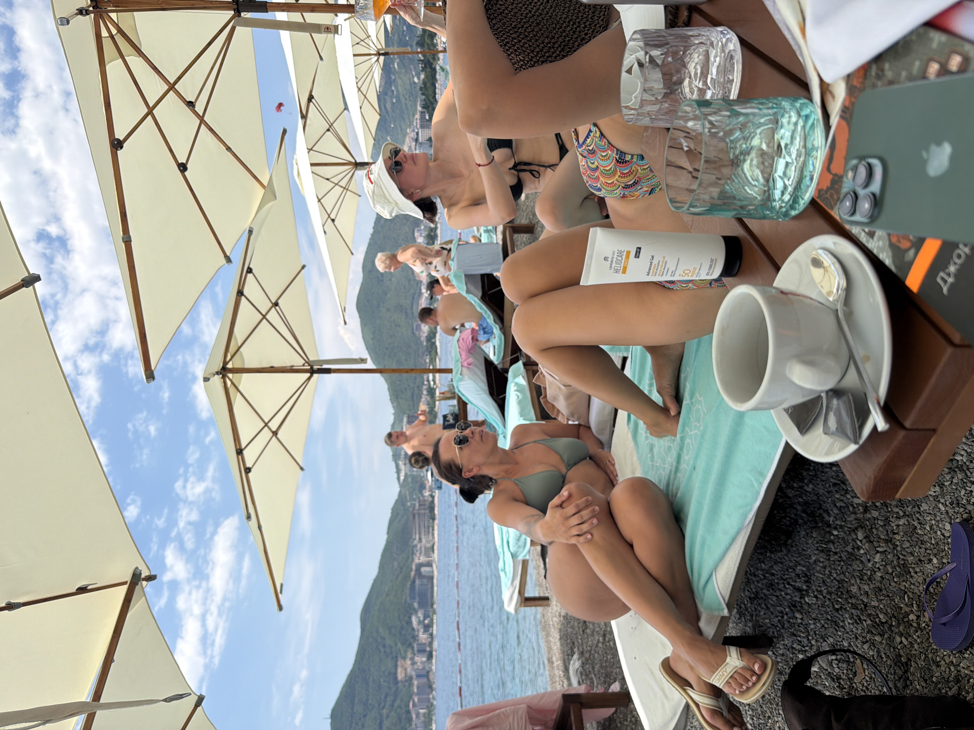 A group of people are enjoying a sunny day at a beach resort in Budva, Montenegro. They are seated on lounge chairs under large, cream-colored umbrellas, with a view of the Adriatic Sea and a lush, mountainous coastline. The scene suggests a relaxed atmosphere, with some individuals sunbathing and others engaged in conversation.
One woman in the foreground, wearing a green bikini and sunglasses, is posed on a lounge chair, looking out towards the sea. To her right, a tube of "HELIOCARE Advanced Gel SPF 50+" sunscreen is visible, indicating a focus on sun protection. Another woman, further back, is wearing a white hat and sunglasses, also relaxing under an umbrella. Other individuals can be seen in the background, some appearing to be swimming or sunbathing on the beach.
The overall ambiance is one of leisure and vacation. The bright sunshine and clear blue sky suggest pleasant weather. The presence of lounge chairs, umbrellas, and the sea indicate a beach resort setting. The image captures a moment of relaxation and enjoyment in a scenic coastal location.