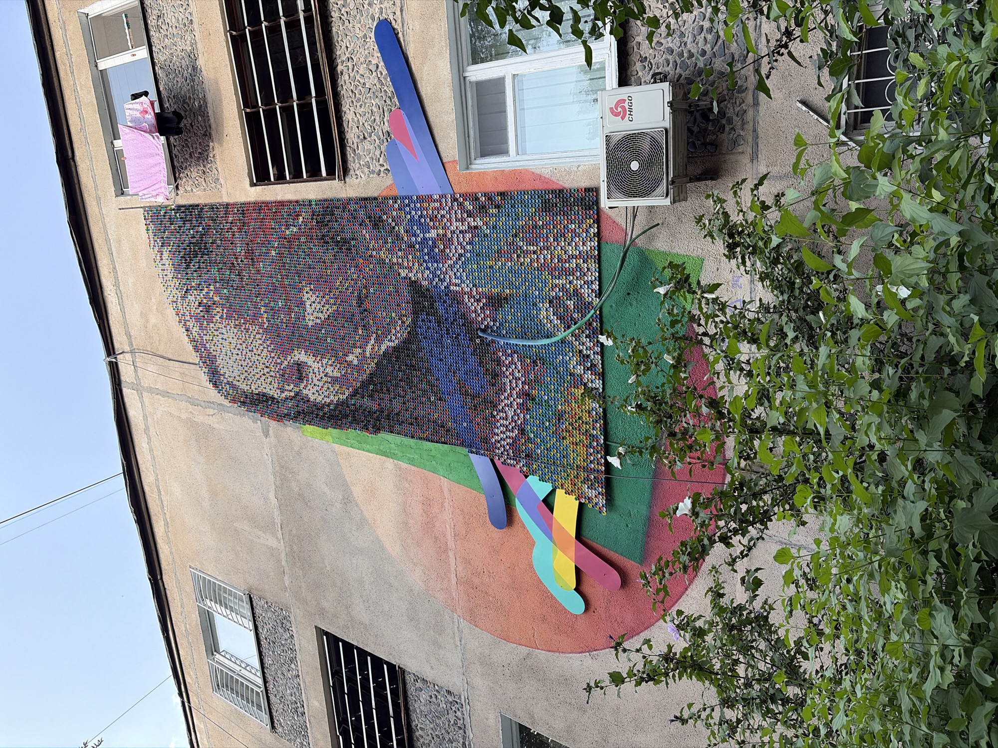 In an urban environment, the side of a multi-story building is adorned with a large mural. The mural features a mosaic portrait of a person's face, composed of numerous small, colorful bottle caps. Abstract shapes and lines in vibrant hues, including pink, orange, yellow, green, blue, and purple, are painted onto the building's facade, creating a dynamic backdrop for the mosaic. A green hose snakes across the mural and down towards the bottom of the frame.
On the building's wall, several windows are visible. One upper-floor window has laundry, including a pink towel and dark clothing, hanging out to dry. Another window on the same level features horizontal blinds. A window on a lower floor is partially obscured by lush green foliage in the foreground. An air conditioning unit is mounted beneath this lower window, bearing the "CHIGO" logo. The building's exterior is a mix of concrete and stone-like textured surfaces. The sky above is a pale blue, with thin overhead wires crossing the frame. The scene is a blend of everyday urban life and artistic expression.