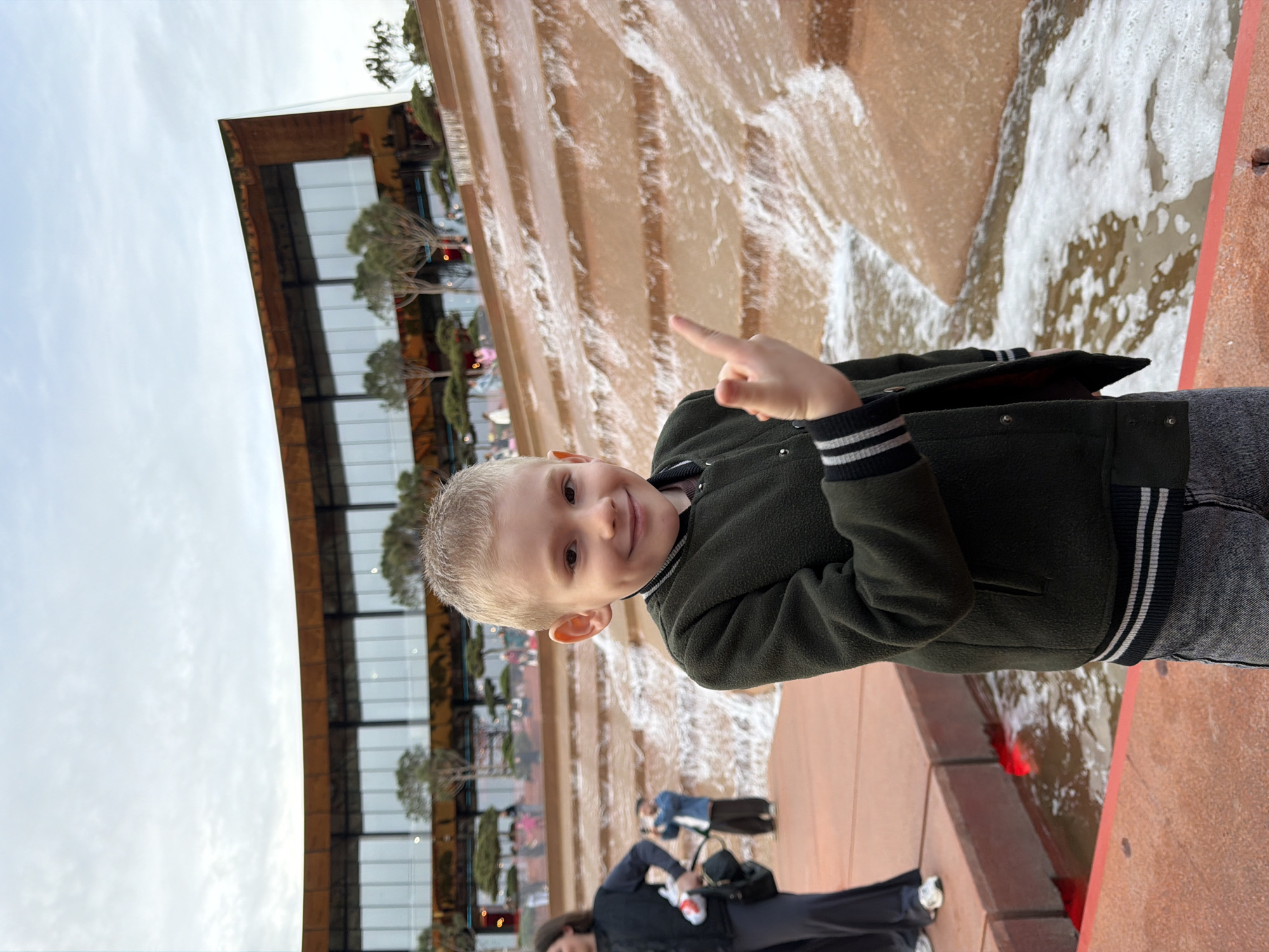 A young boy with blonde hair is posing for a picture in front of the Krasnodar Park in Krasnodar, Russia. The boy is wearing a dark green jacket with white stripes on the sleeves and waist. He is pointing his index finger upwards. Behind him is a tiered waterfall feature with a building visible in the background, reflecting in the waterfall. Several people are walking in the background. The sky is overcast.
