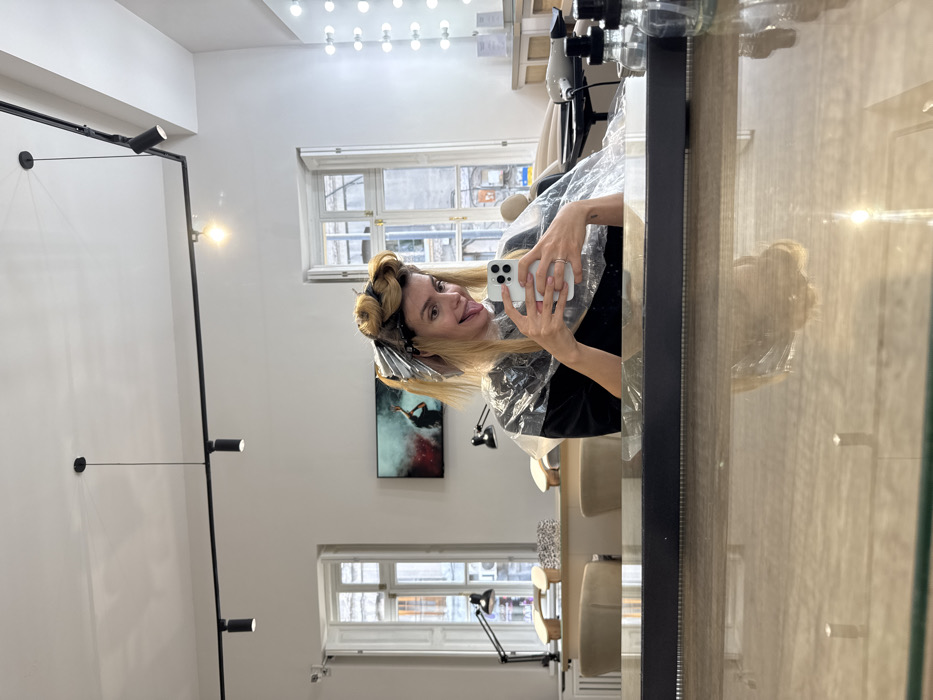 A female individual is captured in a selfie reflected in a mirror within a brightly lit hair and nail salon, located in Tbilisi, Georgia. The specific street location within Tbilisi is not discernible from the image. The subject, a light-skinned woman with blonde hair, appears to be in her 30s. She is smiling and playfully sticking her tongue out while wearing a black top covered by a transparent plastic styling cape. Her hair is undergoing a treatment, with sections wrapped in metallic foils for coloring and other sections rolled into large curlers on top of her head. She holds a white smartphone with a multi-lens camera array in both hands, positioned to take a self-portrait. A simple band-style ring is visible on her left ring finger, and her fingernails are manicured with light pink polish.
The salon's interior features white walls and a white ceiling, which supports a black track lighting system with multiple spotlights and a single black pendant light. To the left, a manicure station is visible, consisting of a long, light-colored wooden table with two beige upholstered chairs, two black adjustable desk lamps, and several wooden hand rests. In the background, two tall, white-framed, multi-paned windows reveal an urban street scene with older stone buildings. Between the windows, a flat-screen television is mounted on the wall, displaying an abstract image of a dark figure against a backdrop of colorful smoke. To the right, a styling station features a large, vertically oriented mirror framed by numerous exposed spherical lightbulbs, with a hairdryer visible on its light wooden counter. The foreground depicts a reflective counter surface, reflecting the woman and overhead lights, upon which two clear glass bottles with black pump tops are positioned.