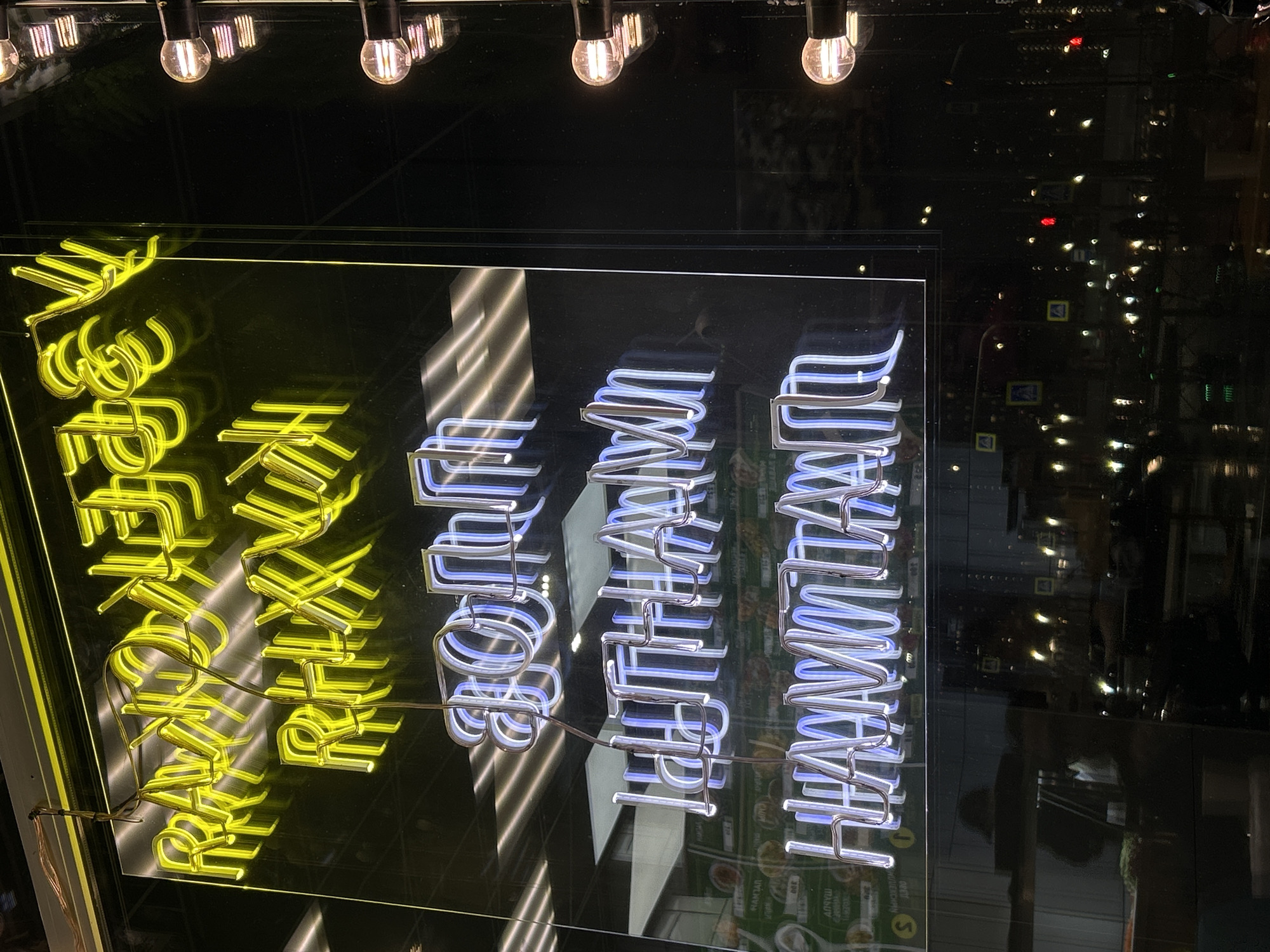 The image, correctly oriented, depicts a nighttime urban scene viewed through a reflective window, likely that of a commercial establishment in Saint Petersburg, Russia. The primary focus is a large, illuminated neon sign against a dark interior background.
The neon sign is composed of two distinct parts. On the left, vibrant yellow neon letters spell out "ХЛЕБ И ЗРЕЛИЩ" (Khleb i Zrelishch), which translates from Russian as "Bread and Circuses" or "Bread and Spectacles." On the right, bright blue neon forms the words "БЫТЬ ДОБРУ!" (Byt' Dobru!), meaning "May good prevail!" or "Let there be good!". The intricate bends of the neon tubes and their support structures are clearly visible, especially for the yellow letters.
The window's surface reflects the external environment, revealing a plethora of distant lights from other buildings and streetlights, typical of a city at night. Among these reflections, several blurry yellow square traffic signs with black arrow symbols can be discerned. Faint reflections of internal elements, possibly menu boards or decor from behind the viewer, are also visible at the bottom right. Here, some very blurry Cyrillic text like "РАМТАЙ" and "АЙРУШ" is faintly legible, alongside circular yellow symbols containing numbers '5' and '1', suggesting the establishment might be a restaurant or café. Above the neon sign, several exposed incandescent lightbulbs hang, casting warm, circular glows and adding to the ambient lighting. The scene captures a moment of urban vibrancy, illuminated by neon and reflections.
