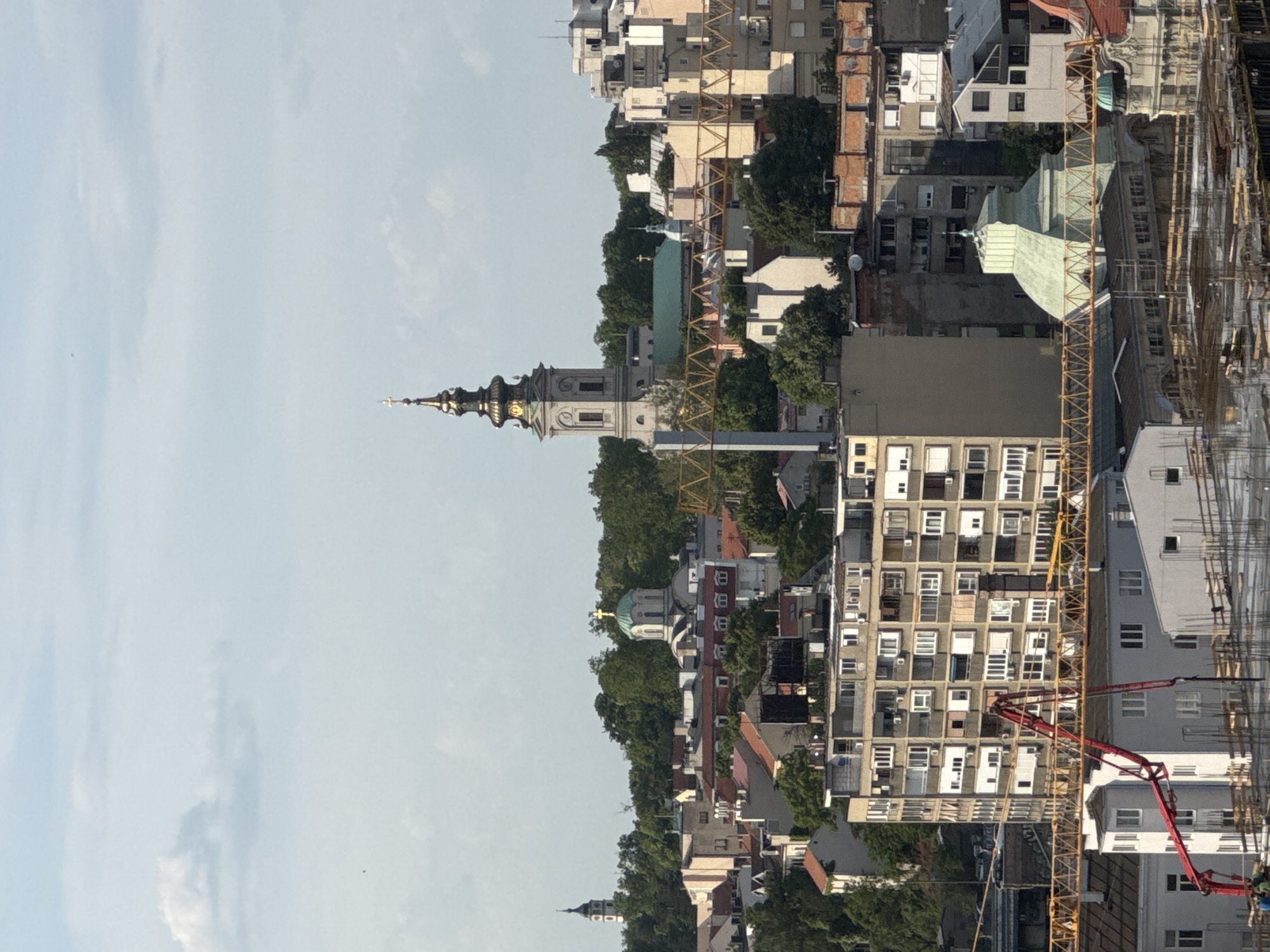 This aerial view shows a section of Belgrade, Serbia. The sky is partly cloudy. A church with a distinctive spire is visible on the left, likely the Saborna Church. Numerous buildings of varying heights and architectural styles dominate the scene, including residential buildings and structures under construction, indicated by cranes. Green trees are interspersed among the buildings. A pumping truck is shown on the right.