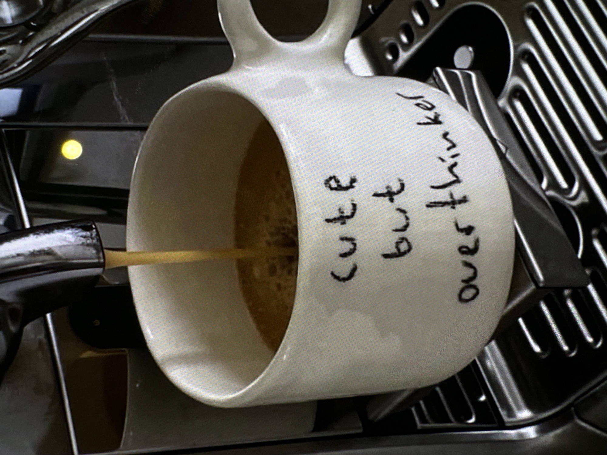 A close-up shot shows a white mug with the words "cute but overthinker" written on its side, being filled with coffee from an espresso machine. The mug is placed on the metal drip tray of the machine. The background consists of the machine's metal components, with a small yellow light visible on its side. There is no indication of the location being Yerevan, Armenia.