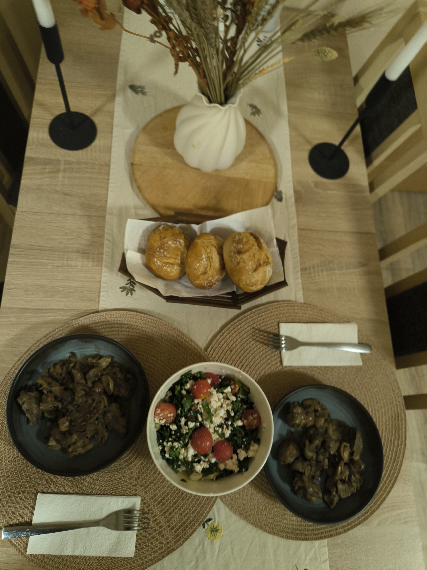 An overhead view of a dining table set for a meal in Unknown, Unknown. The light-colored wooden table features a central off-white table runner adorned with dark brown stylized leaf patterns. At the top center, a round wooden board supports a white pumpkin-shaped candle. Behind it, a white, ribbed vase holds a bouquet of dried grasses, wheat stalks, and orange-brown autumn leaves. Two black metal candlesticks with white taper candles are visible on the table surface, one at the far left and one at the far right.
A small, rectangular wooden tray contains three golden-brown bread rolls, resting on a white paper napkin or towel. Below this, three round, woven placemats are visible. On the left placemat, a dark blue-grey plate holds a portion of a dark brown, rich-looking stewed dish, likely consisting of meat (such as chicken gizzards or liver) and mushrooms. A silver fork with a textured handle rests on a white paper napkin positioned below this plate.
In the center foreground, a white bowl contains a colorful salad composed of dark green leafy greens (possibly kale or spinach), crumbled white cheese (feta), and halved red cherry tomatoes. On the right placemat, a matching dark blue-grey plate holds a similar portion of the stewed dish. A silver fork with a smooth handle rests on a white paper napkin positioned above this plate. No individuals are present in the image.