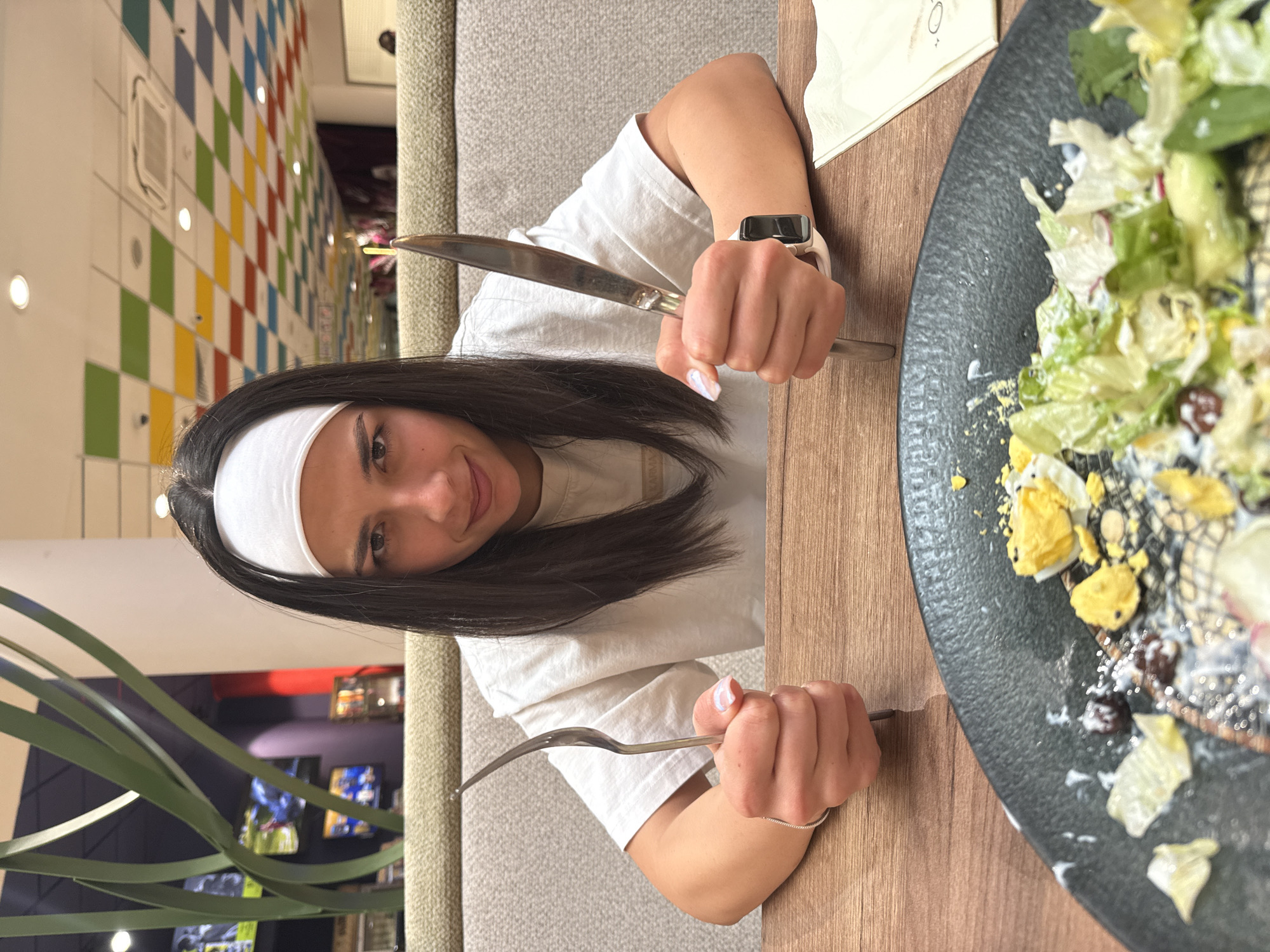 The image captures a woman seated at a wooden table in an indoor dining establishment, identified as being in Veliko Tarnovo, Bulgaria. The woman, with long dark hair and a white fabric headband, looks directly at the camera with a slight smirk. She is wearing a light-colored, short-sleeved t-shirt, with faint text visible on her right shoulder. In her left hand, she holds a large fork vertically, and in her right hand, she holds a large knife, also positioned vertically. A thin silver chain bracelet is visible on her left wrist, and a light-colored smartwatch with a dark screen is on her right wrist. Her fingernails have a light-colored, possibly iridescent, polish.
On the wooden table in front of her is a dark, textured plate containing a partially consumed salad. The salad ingredients visible include green lettuce leaves, sliced hard-boiled eggs, and small dark components, possibly olives or berries, mixed with a light-colored dressing. A white paper napkin is visible to the right of the plate.
Behind the woman, a beige upholstered booth-style seating extends across the mid-ground. To the left of the woman, large, abstract green sculptural elements are visible, along with multiple wall-mounted screens displaying various media, suggesting a recreational or entertainment area. The wall behind these elements appears dark purple. To the right of the woman, the ceiling features a grid pattern of colorful square panels in red, yellow, green, and blue, interspersed with white ceiling tiles containing recessed lighting fixtures and an air conditioning vent.