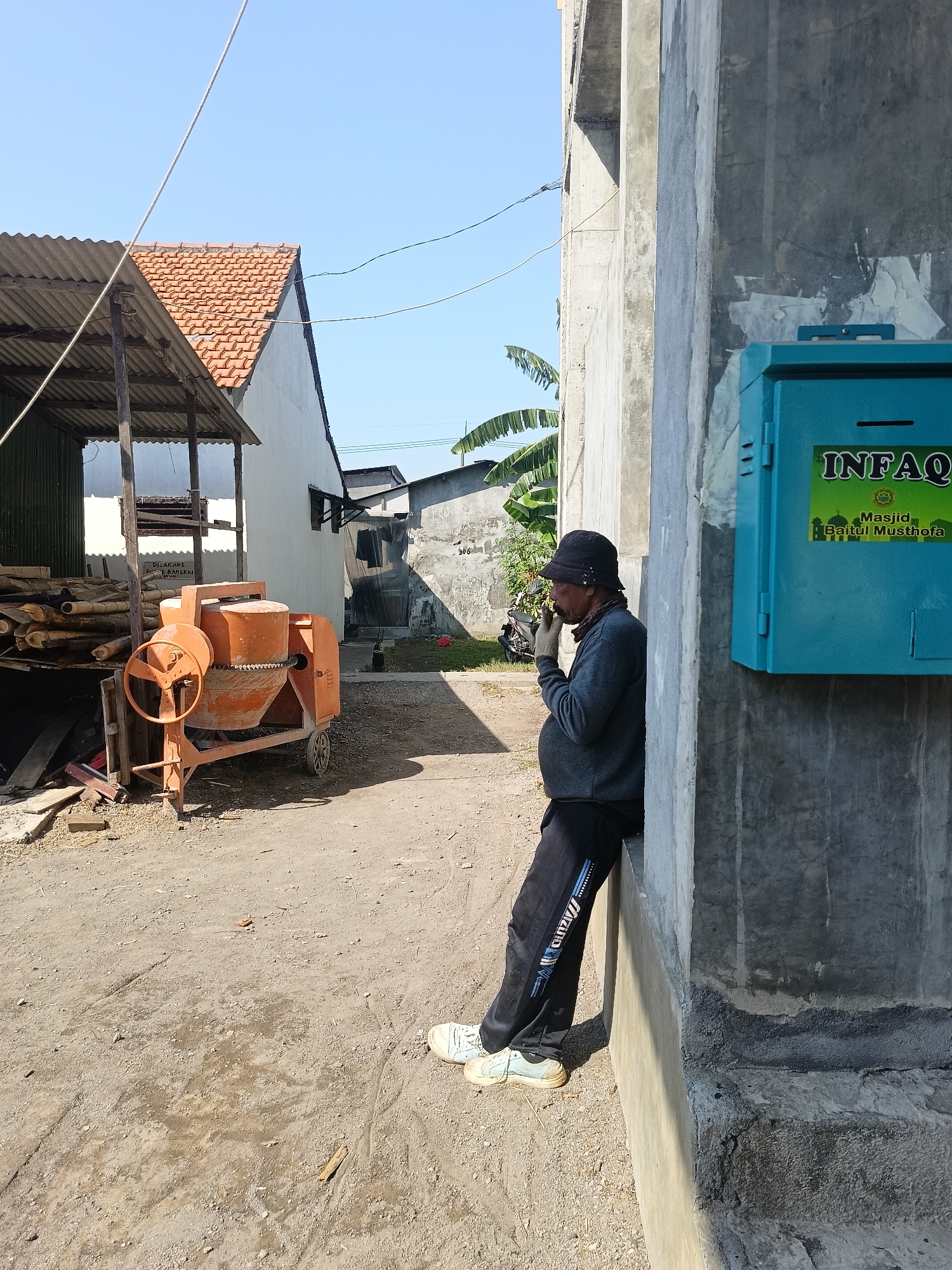 A man wearing a black bucket hat, a grey long-sleeved shirt, black pants, and white sneakers is leaning against a concrete wall and smoking. He has a grey bandana tied around his neck. In the background, there is an orange cement mixer, a pile of wood, and a building with a red tile roof. A blue donation box labeled "INFOQ Masjid Baitul Musthofa" is attached to the concrete wall next to the man. The scene appears to be an outdoor work or construction area in the city of Gedangan, Indonesia, under a clear blue sky.