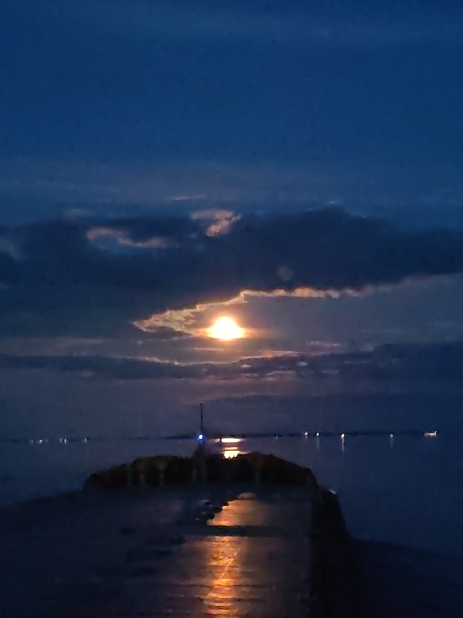 The image captures a serene, dark waterscape at night or during deep twilight in Bol'shie Mkhi, Russia. The dominant feature is a brilliant, intense celestial body, likely the moon, positioned centrally in the upper half of the frame. It glows with a bright orange-yellow light, partially obscured by dark, dense clouds that stretch across the sky. Patches of lighter cloud are illuminated around the moon, creating a dramatic halo effect.
In the foreground, a dark, solid structure, possibly a pier or a breakwater, extends from the bottom center into the body of water. Its wet surface reflects the intense light source above, creating a distinct, shimmering orange-yellow streak that leads towards the viewer. On this structure, near the middle distance, a faint, small blue light can be seen on a slender pole-like object, possibly a navigation aid or part of a small beacon.
The water, which occupies the lower half of the frame and extends into the distance, appears calm and dark, reflecting hints of the dim light from the sky. Across the distant horizon, several small, scattered white lights are visible on the water, suggesting the presence of distant boats, buoys, or shore lights. No people are visible in the scene. The overall ambiance is tranquil and contemplative, with the bright moon serving as the focal point against the dark, cloudy sky and water. No visible text is present.