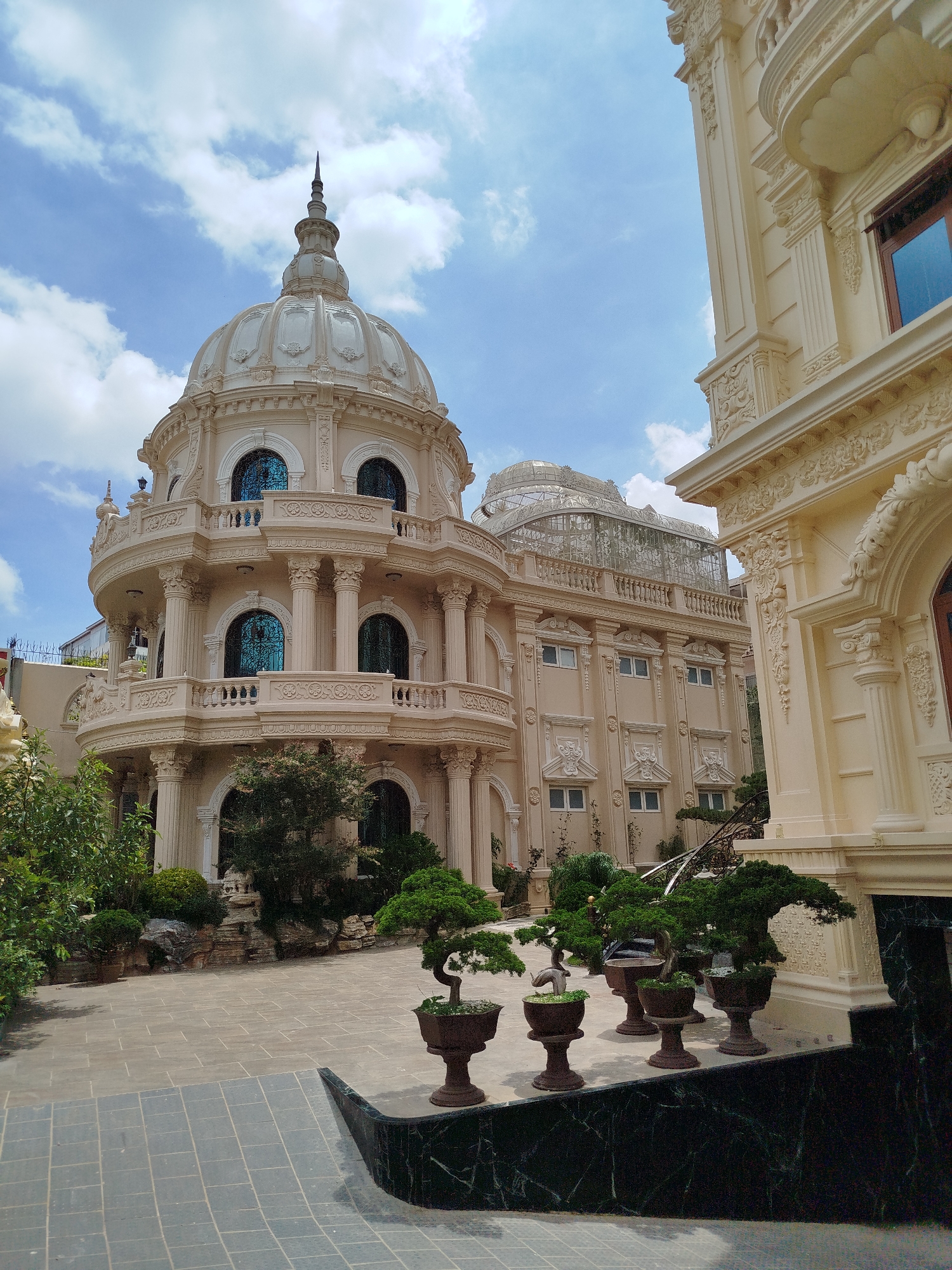 The image shows a large, ornate, cream-colored building with a prominent dome, reminiscent of a European palace or temple. The architecture features classical columns, balconies with intricate railings, and decorative carvings. The building appears to be a religious site or a grand residence. In the foreground, there is a courtyard paved with stone tiles, and several potted bonsai trees are arranged along a dark green marble ledge. Lush greenery and trees surround the building. The sky is blue with scattered white clouds. This building is the Huyen Tran Cong Chua Temple in Da Lat, Vietnam. There are no people visible in the image.