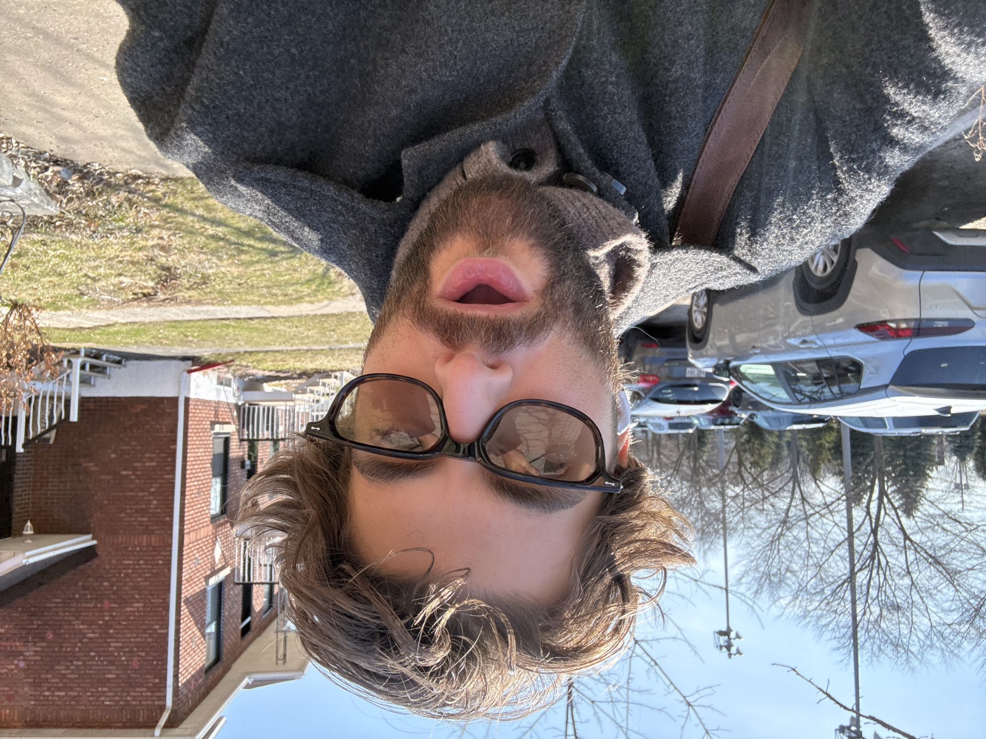A close-up selfie of a person with brown hair, a beard, and glasses, wearing a gray coat and a brown strap across the chest. The background includes a brick building, a street, parked cars, and trees. The photo appears to be taken on a sunny day. There is no indication of the location being in Montreal, Canada based on the image.