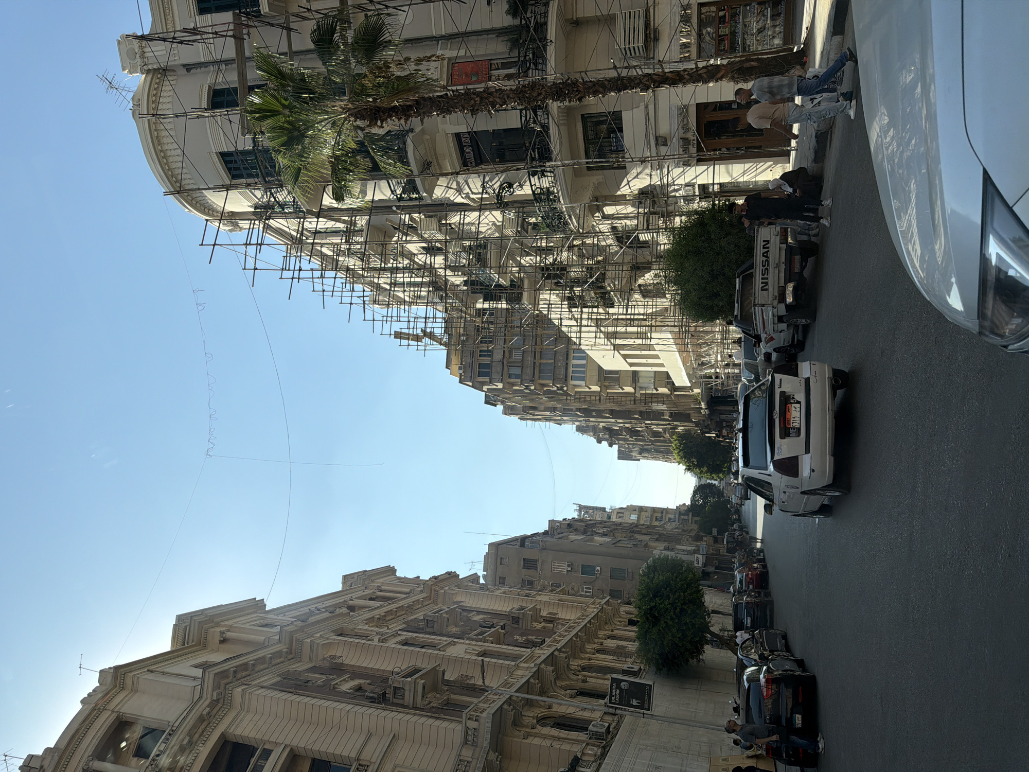 The image depicts a sunlit street scene in Cairo, Egypt, characterized by the presence of multi-story buildings and parked cars. On the left, a grand, ornate building with balconies and intricate architectural details dominates the foreground. Across the street, several buildings are visible, with one on the right side of the image covered in scaffolding, suggesting ongoing construction or renovation. A tall palm tree stands beside this scaffolded building.
The street is paved and appears to be a two-way road. Several cars are parked along the sides of the street, including a white sedan with a license plate visible in the center of the frame, and a black hatchback on the left. A white pickup truck with "NISSAN" branding is parked near the center of the image.
Several individuals are present. On the left sidewalk, a man is leaning against a parked black car. Further down the street, a group of people are standing near the Nissan truck. On the right sidewalk, two men are walking, and another person is seated on the curb. A third person, possibly a woman wearing a headscarf, is also seated on the curb.
The sky is clear and blue, with the sun casting strong shadows, indicating it is likely daytime. Thin wires or cables are strung across the sky between buildings, possibly for lighting or other urban infrastructure. A banner with text, including "DR. AMIRA LASHIN" and what appears to be medical or professional information, is affixed to a wall on the left. Another sign on the right building reads "GAILO INN" above a doorway. The overall atmosphere is that of a busy urban environment with a mix of older and newer structures, and signs of everyday life and development.
