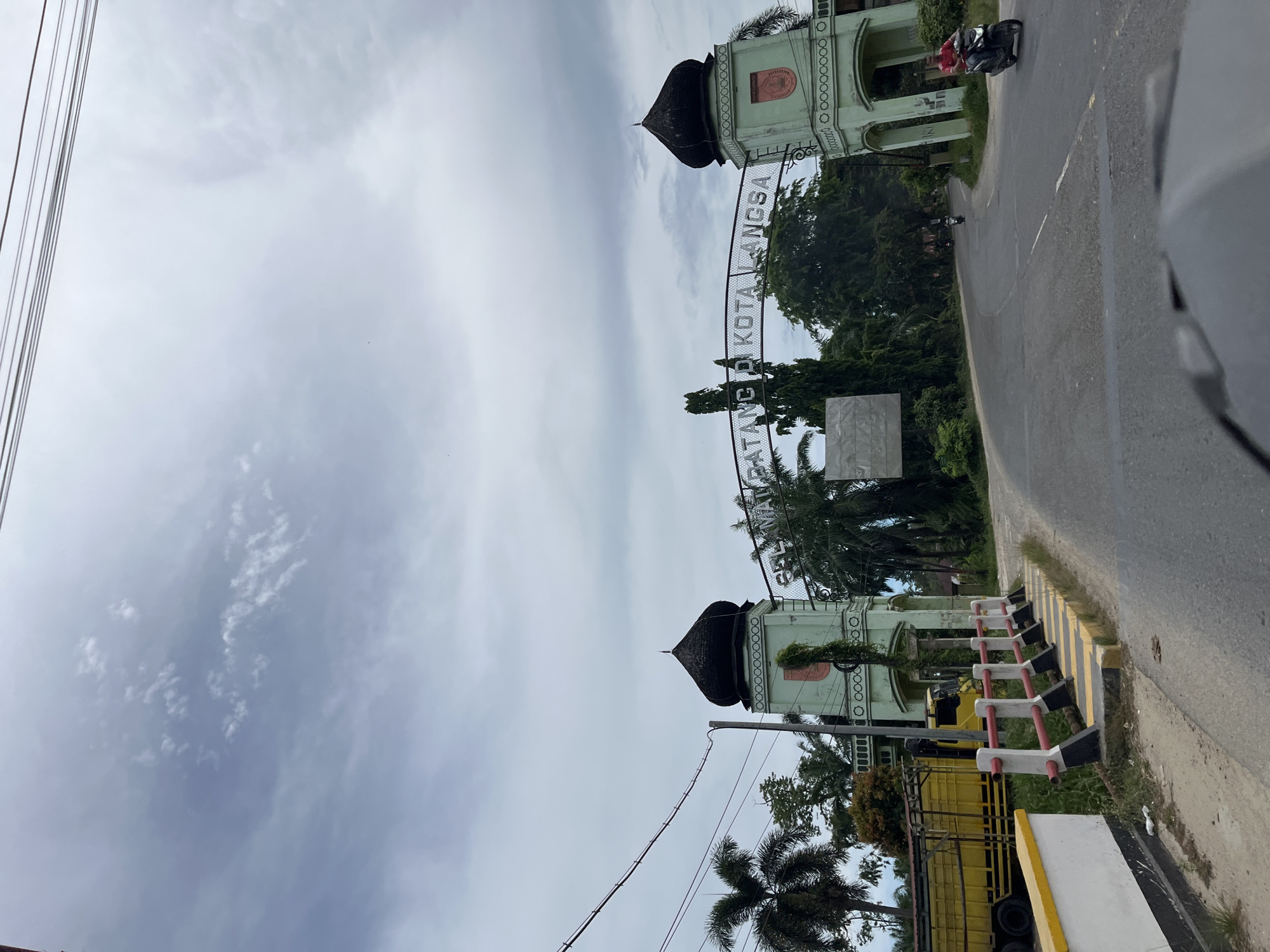 The image depicts an archway marking the entrance to the city of Seuneubokpunti, Indonesia. The arch is adorned with the words "SELAMAT DATANG DI KOTA LANGSA," indicating "Welcome to Langsa City." The archway is flanked by two light green structures topped with dark domes. The left side of the scene features a bright yellow truck parked on the side of the road, adjacent to a white and yellow barrier. Palm trees and other lush greenery surround the archway. The road curves into the distance, where several motorbikes can be seen. The sky above is overcast with visible clouds.